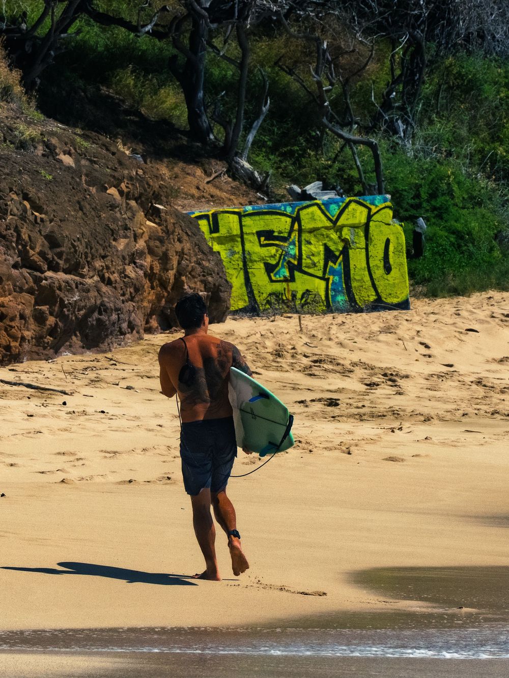 A shirtless surfer carries a green surfboard across the sand at Waikiki toward the water, a graffiti wall and trees along the shore behind.