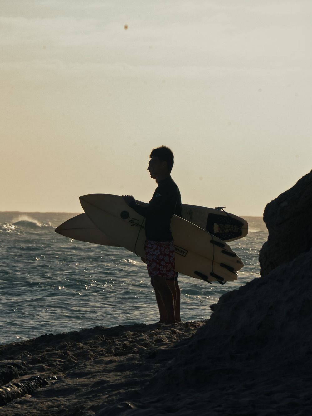 A surfer in red floral boardshorts holds a pale surfboard on a rocky point at Waikiki, watching the waves in soft golden evening light.