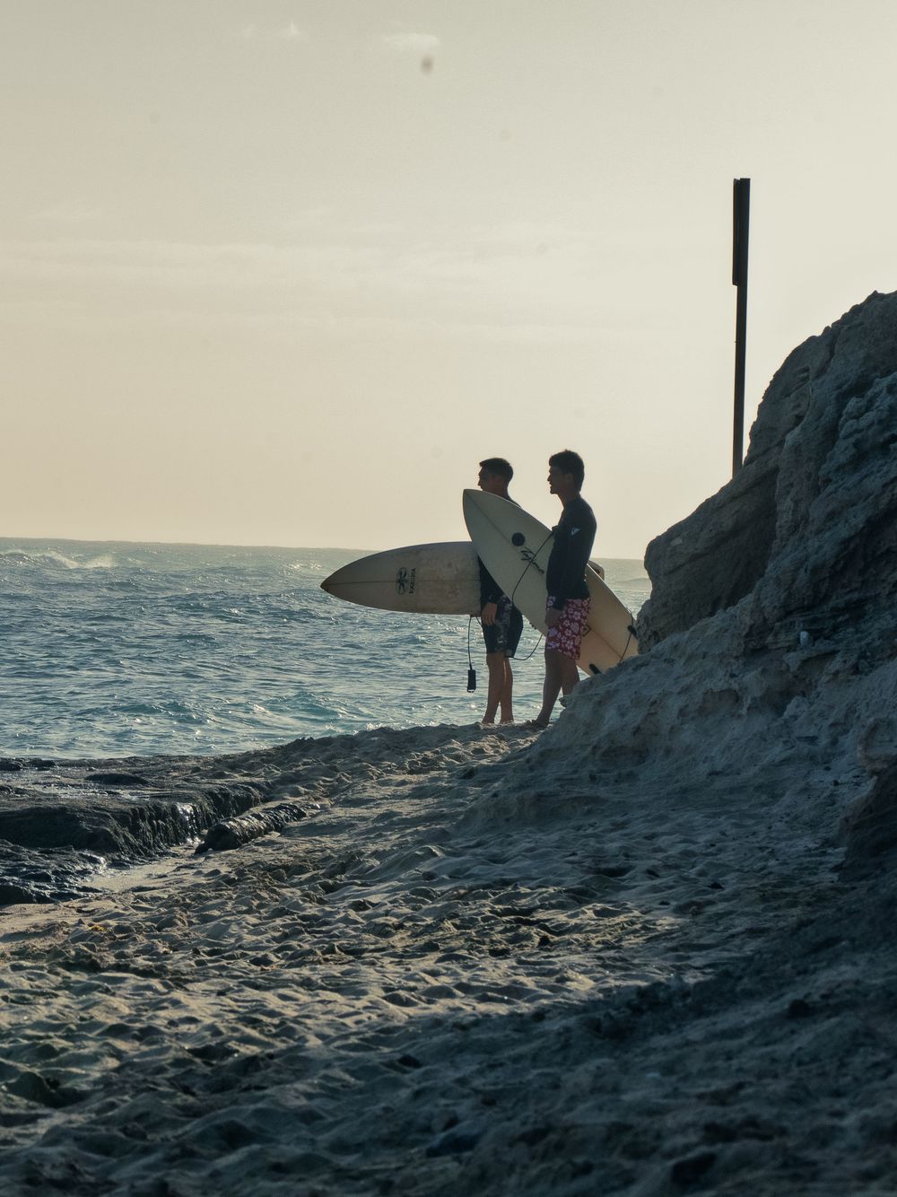 Two surfers stand on a rocky point at Waikiki holding pale surfboards, studying the breaking waves under a hazy sky before paddling out.