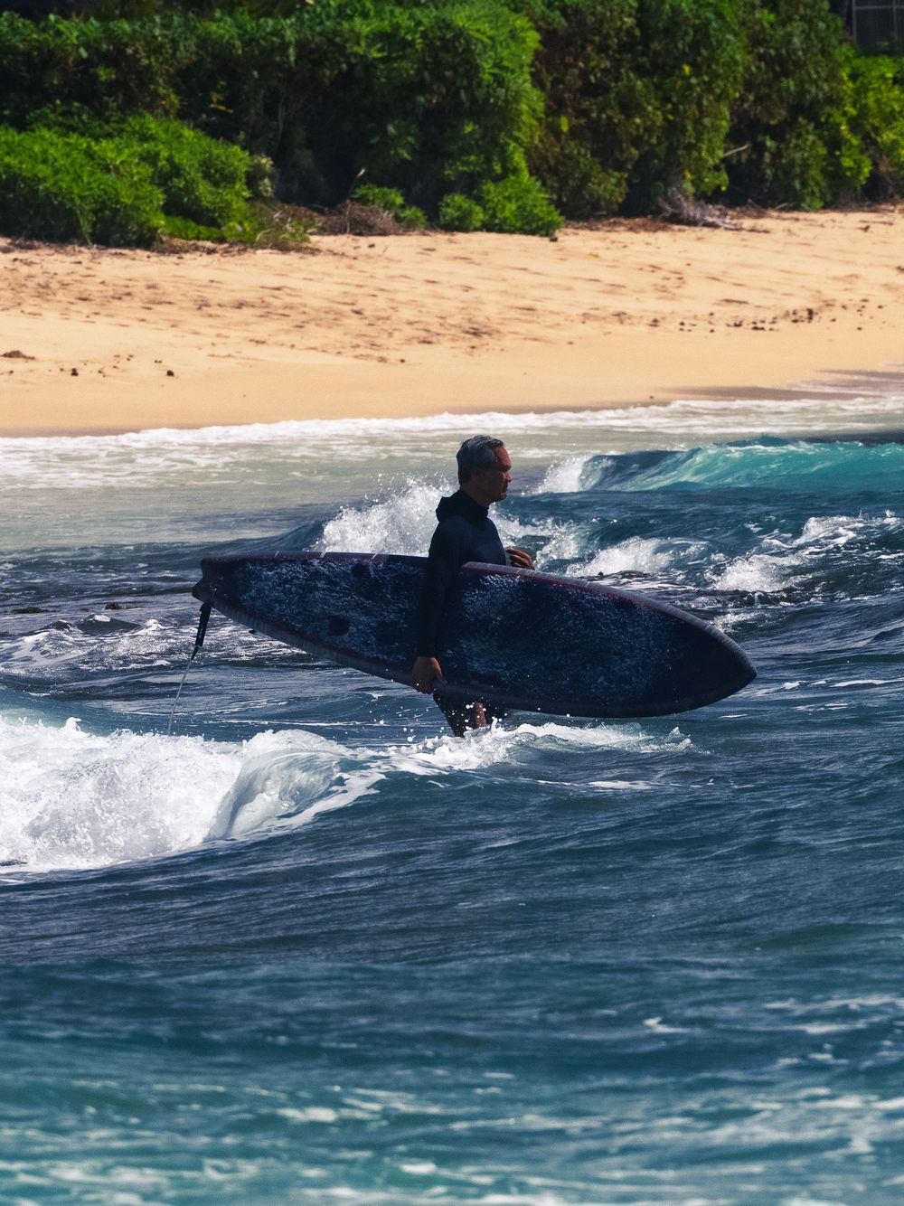 A gray-haired surfer in a black wetsuit wades through the surf at Waikiki carrying a dark surfboard, breaking waves and sand behind him.