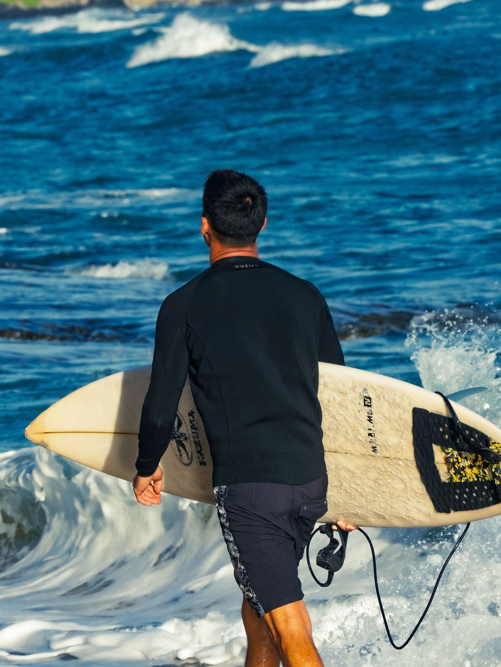 A surfer in a black rash guard walks into the whitewater at Waikiki carrying a pale surfboard under his arm, deep blue ocean ahead.