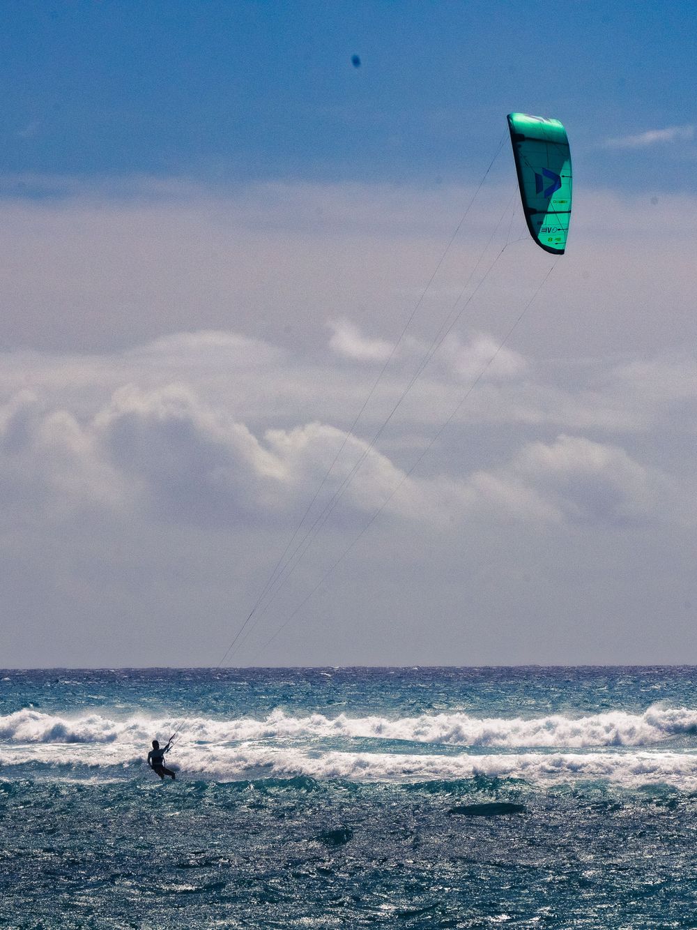 A kitesurfer glides across choppy water at Waikiki, a large teal kite arcing high on long lines against a bright, cloud-streaked sky.