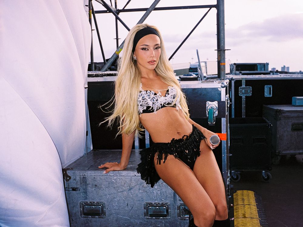 Blonde woman in black fringe shorts and floral crop top posing backstage at an outdoor concert venue.