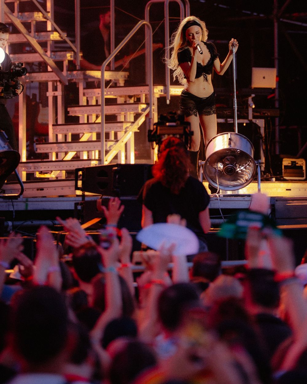 Female performer singing on stage at a packed outdoor concert, with crowd raising hands and phones.
