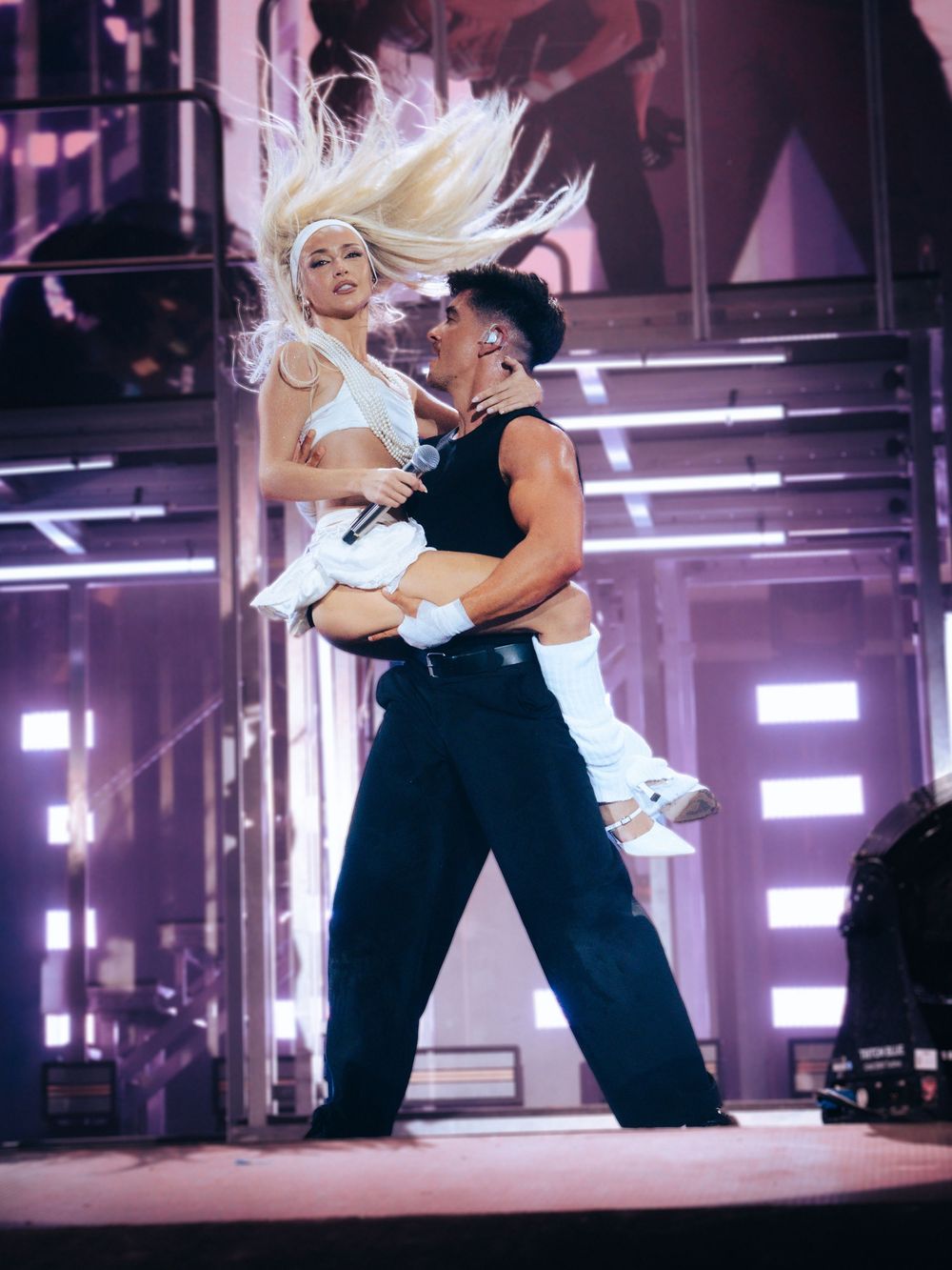 Two performers dance on a lit stage, with a woman in white lifted by a man in black during a live concert.