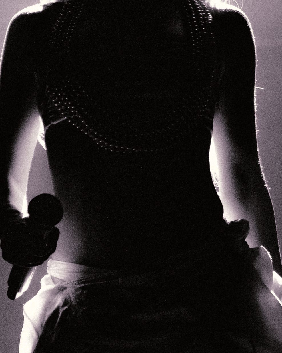 Silhouetted performer holding a microphone on stage, backlit with dramatic lighting highlighting their outfit.