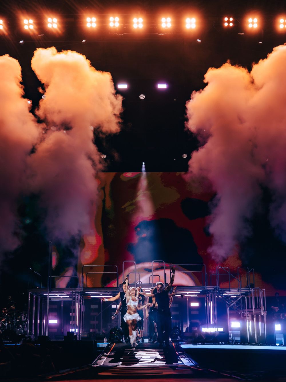 Performers on stage with dramatic pink smoke clouds and colorful lights during a live concert show.