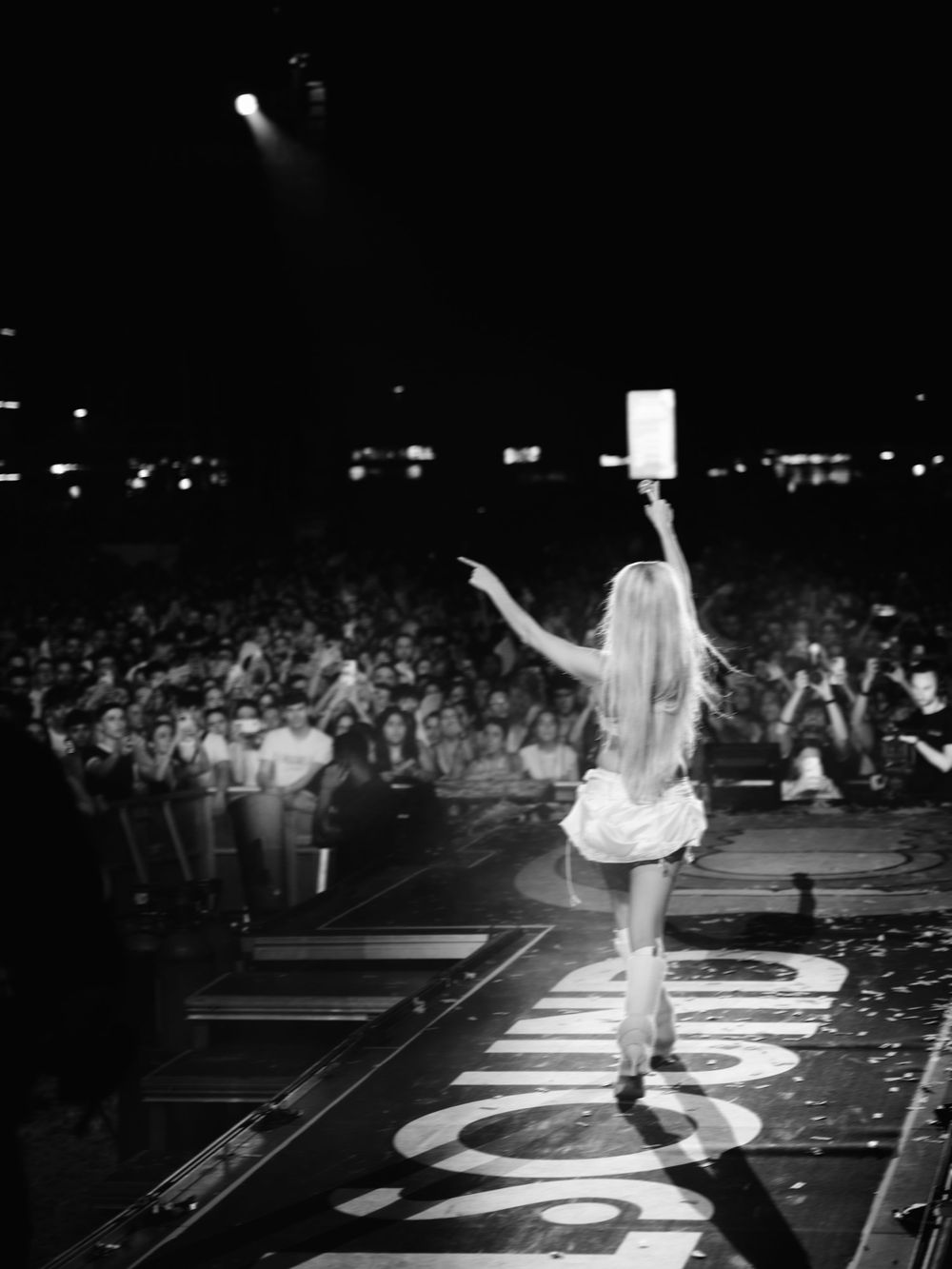 Black and white photo of a female performer on stage holding a sign, pointing at a large concert crowd.