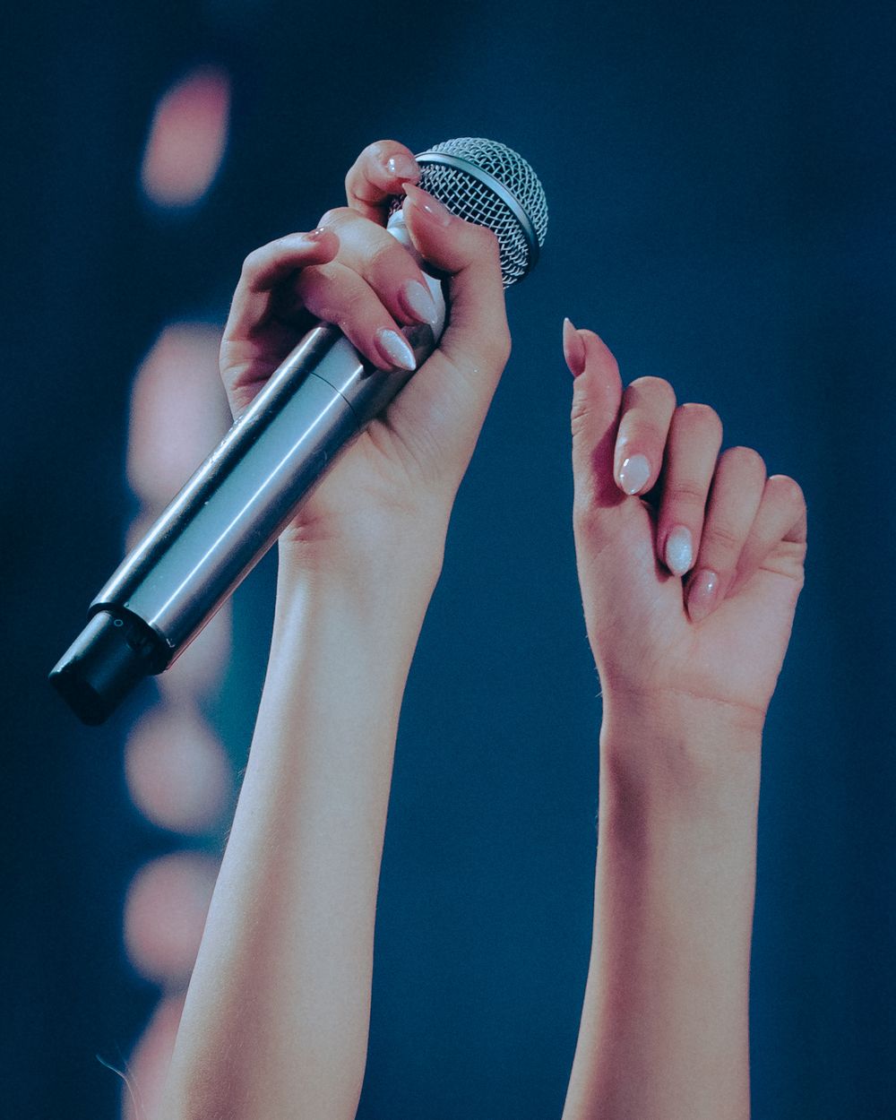 Two hands raised holding a wireless microphone on a blue bokeh stage background.
