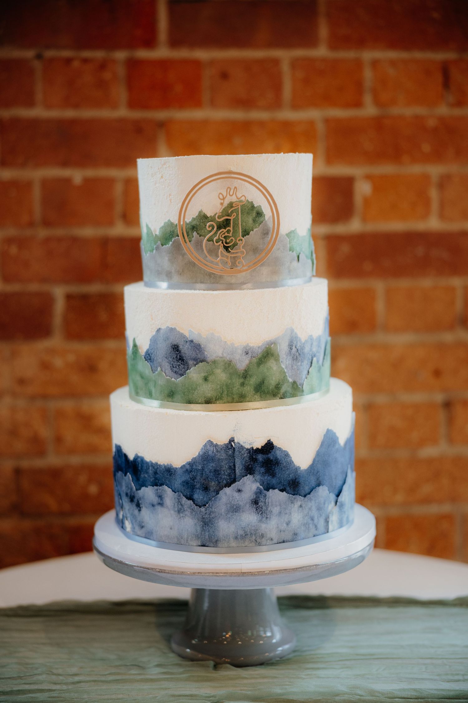 Three-tier white wedding cake with watercolor mountain design in blue and green, gold monogram topper, on gray cake stand.