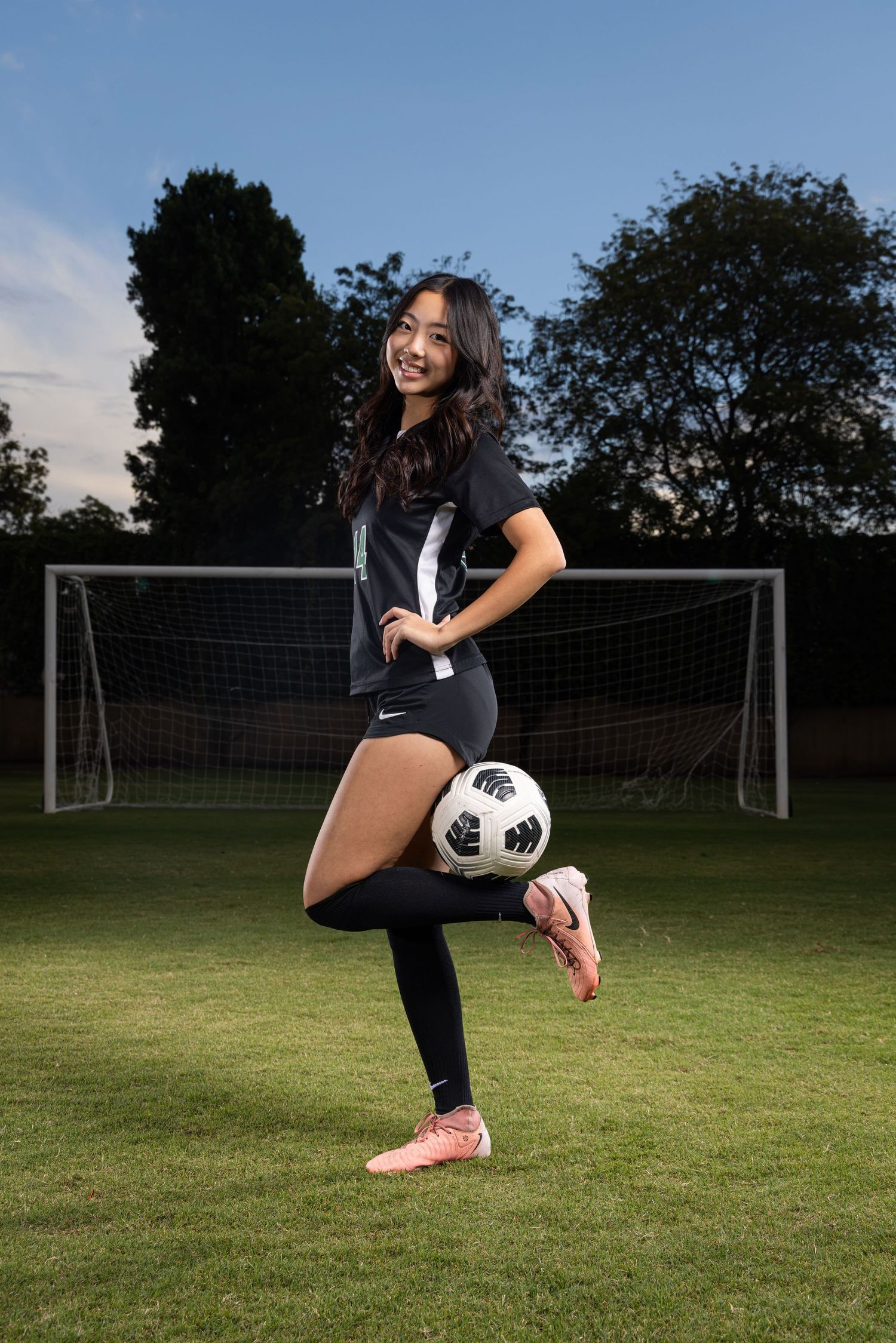 Young female soccer player posing with ball on field, wearing Nike uniform and pink cleats in front of goal net.