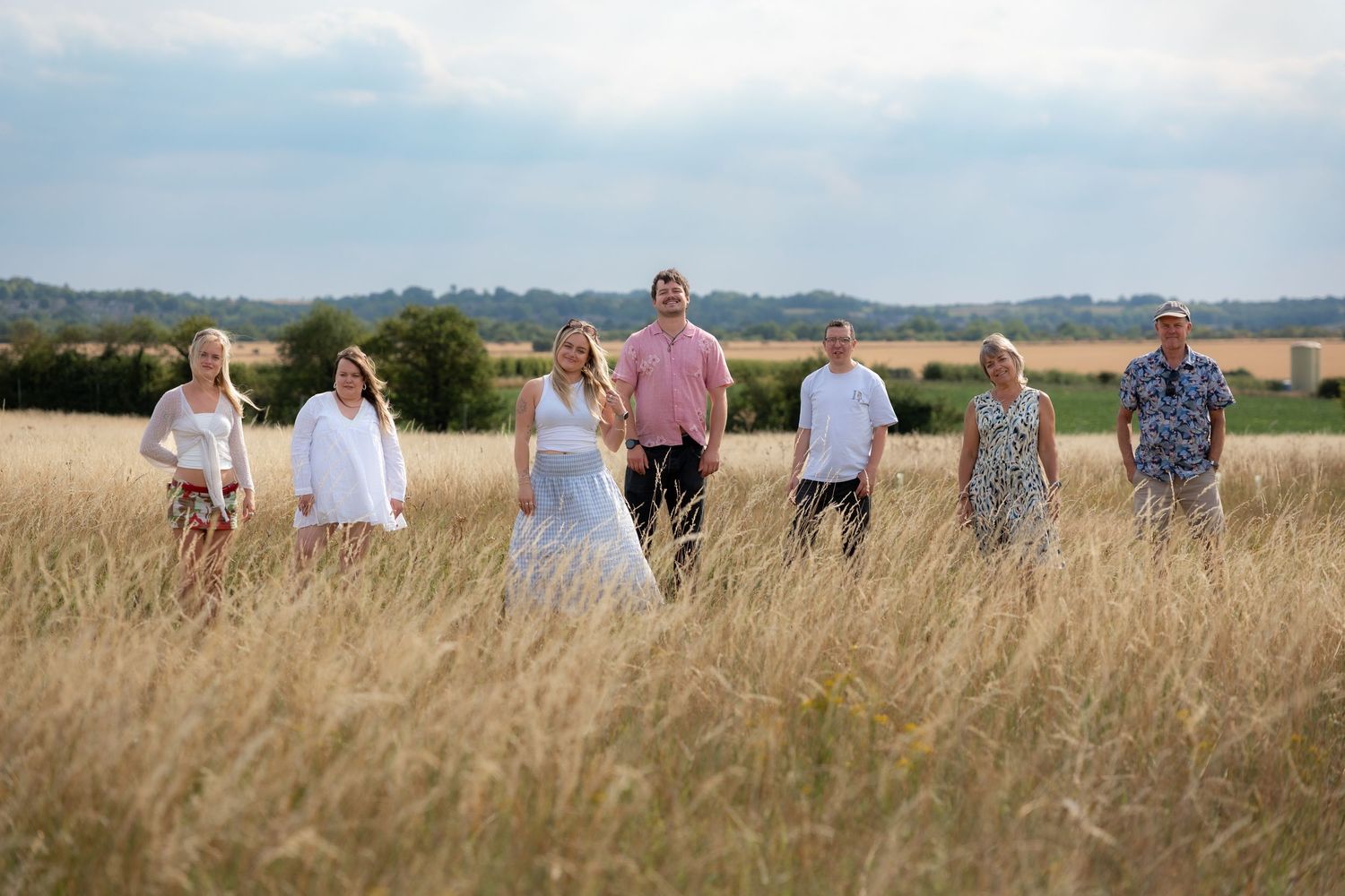 Seven people standing in a golden wheat field on a sunny day with green trees and blue sky in the background.