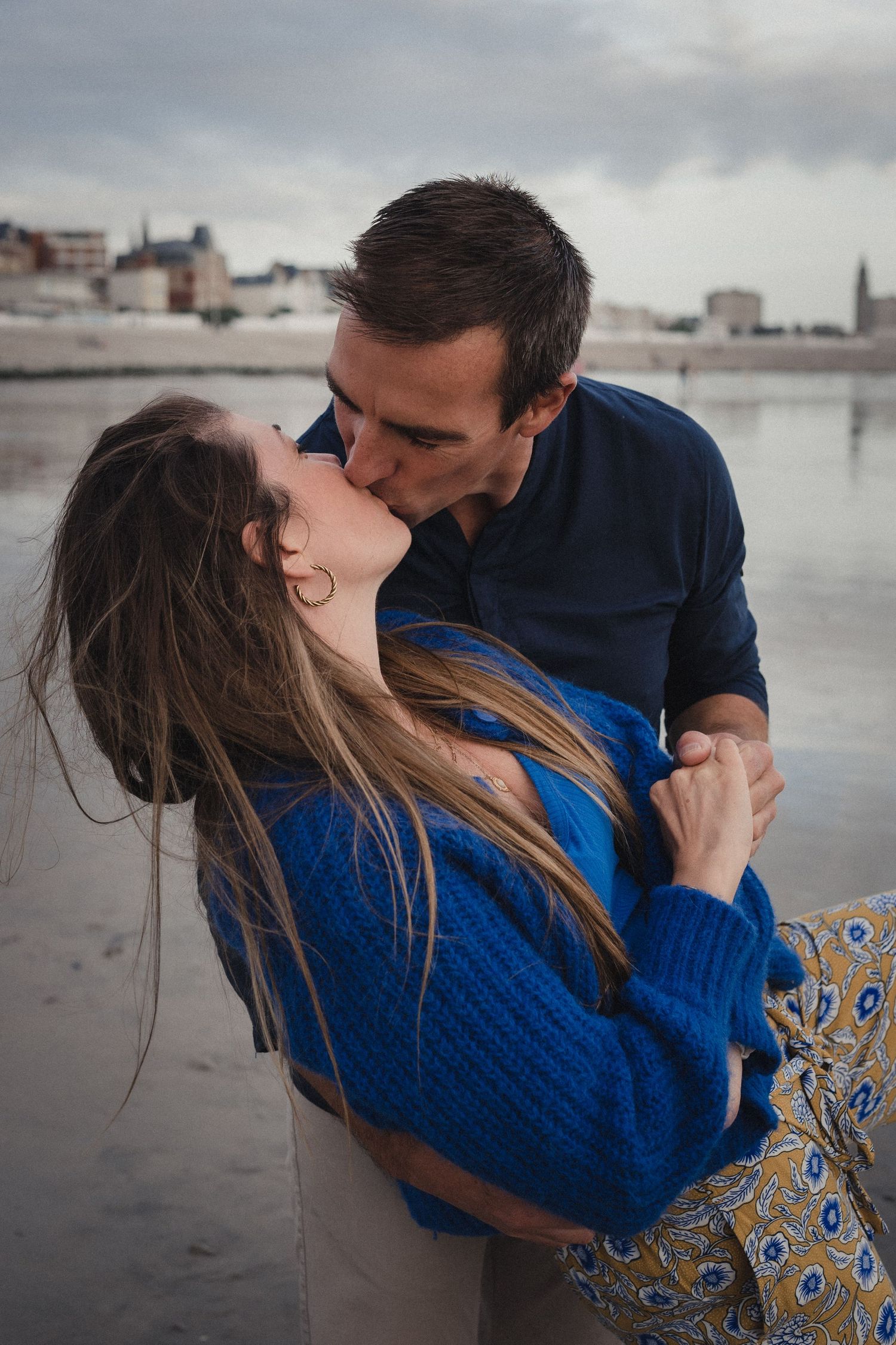 A romantic couple embraces on a cloudy beach with water and buildings visible in the background.