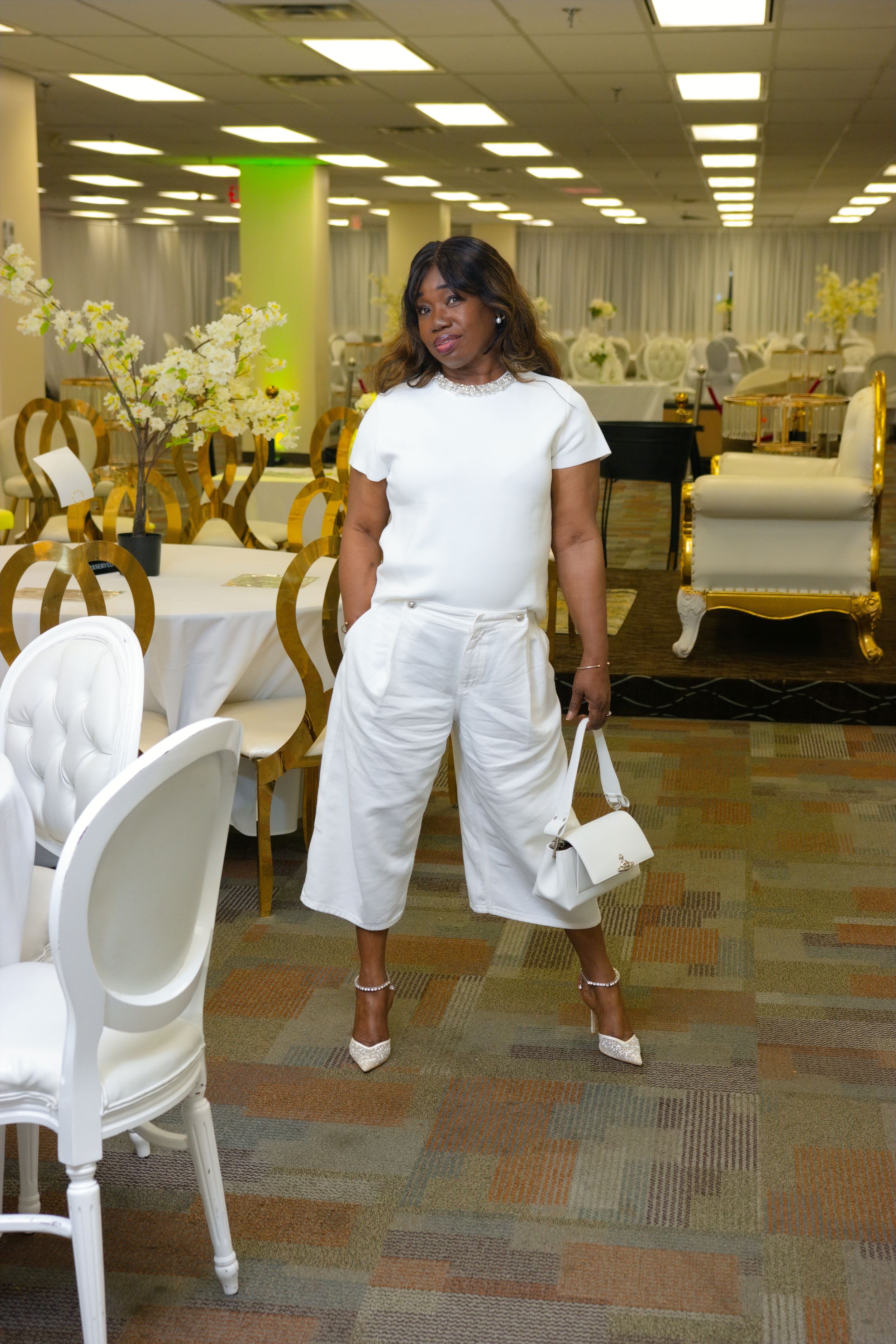 A woman in an all-white outfit with culottes, heels, and handbag poses in an elegantly decorated event venue.