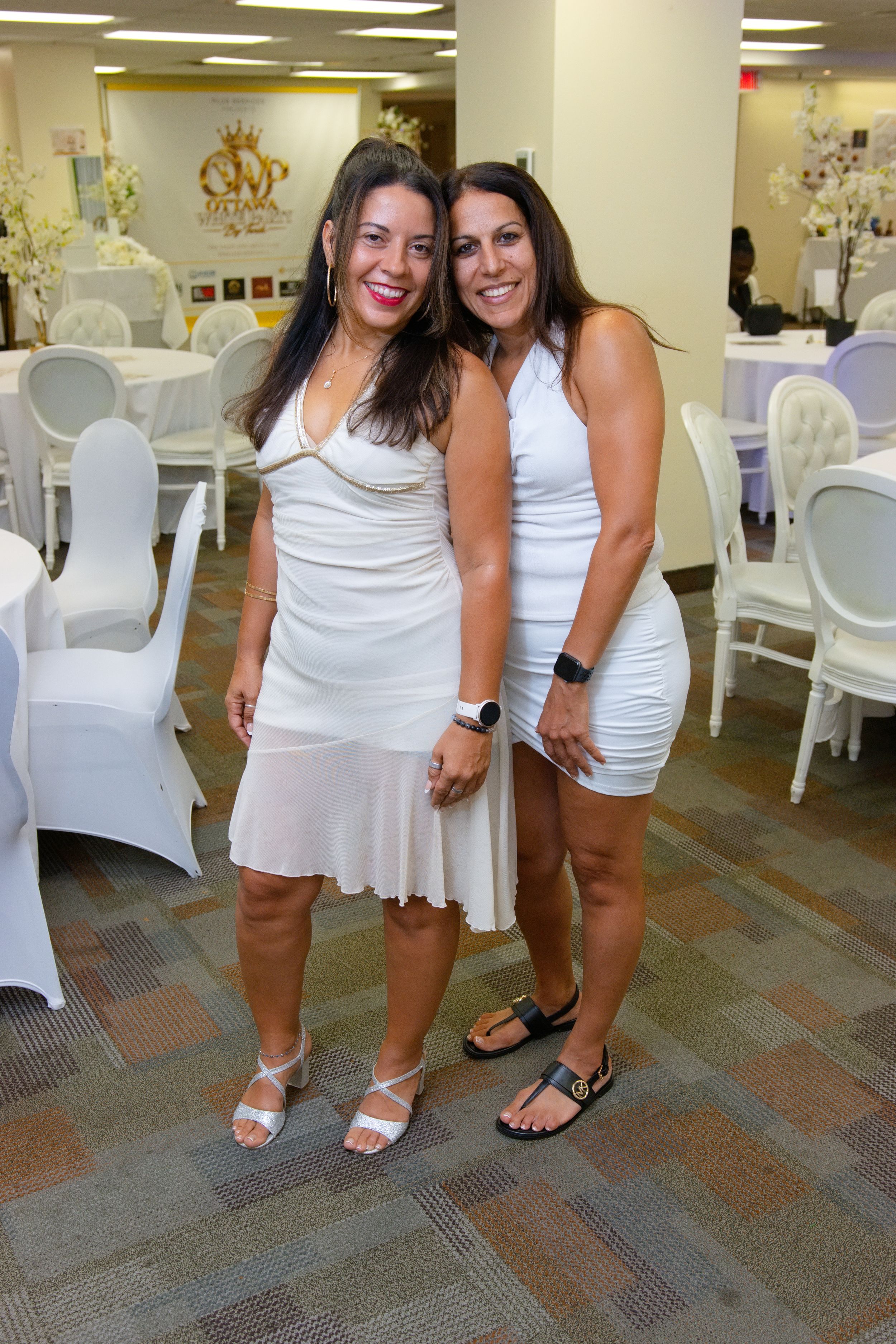 Two women in white dresses smile together at an elegant white-themed event venue.