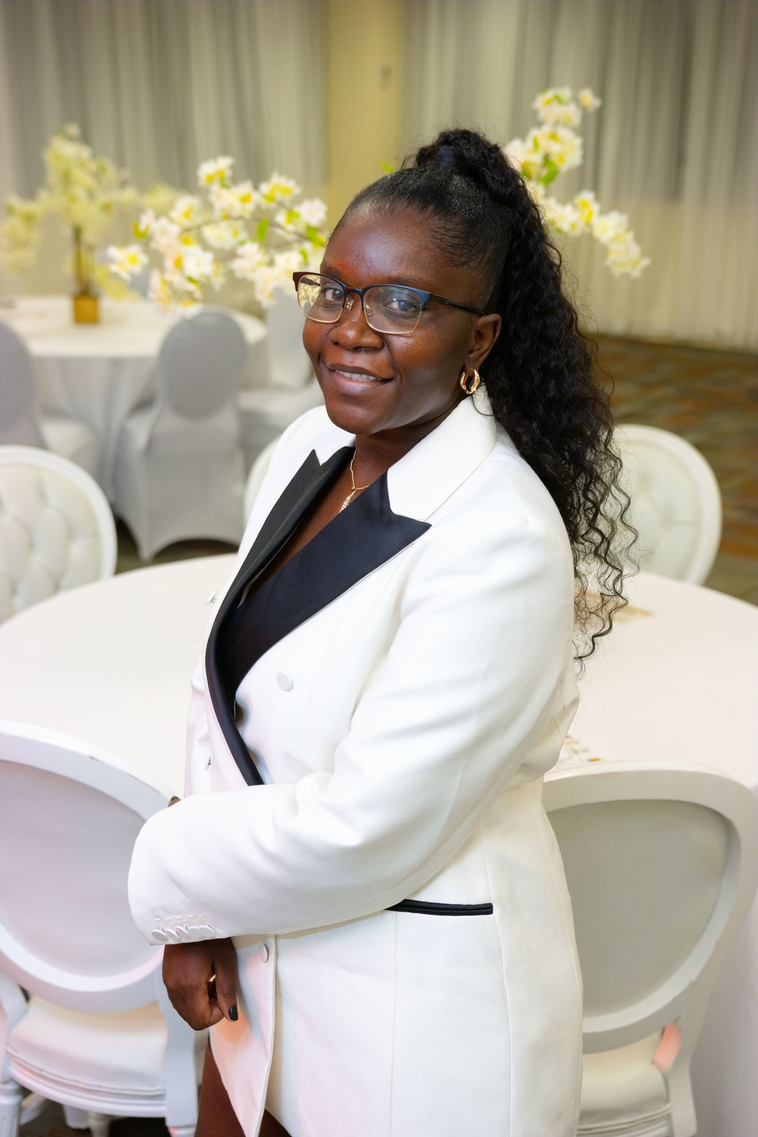 A smiling woman in a white blazer with black lapels poses in an elegant event venue with white chairs and floral decor.