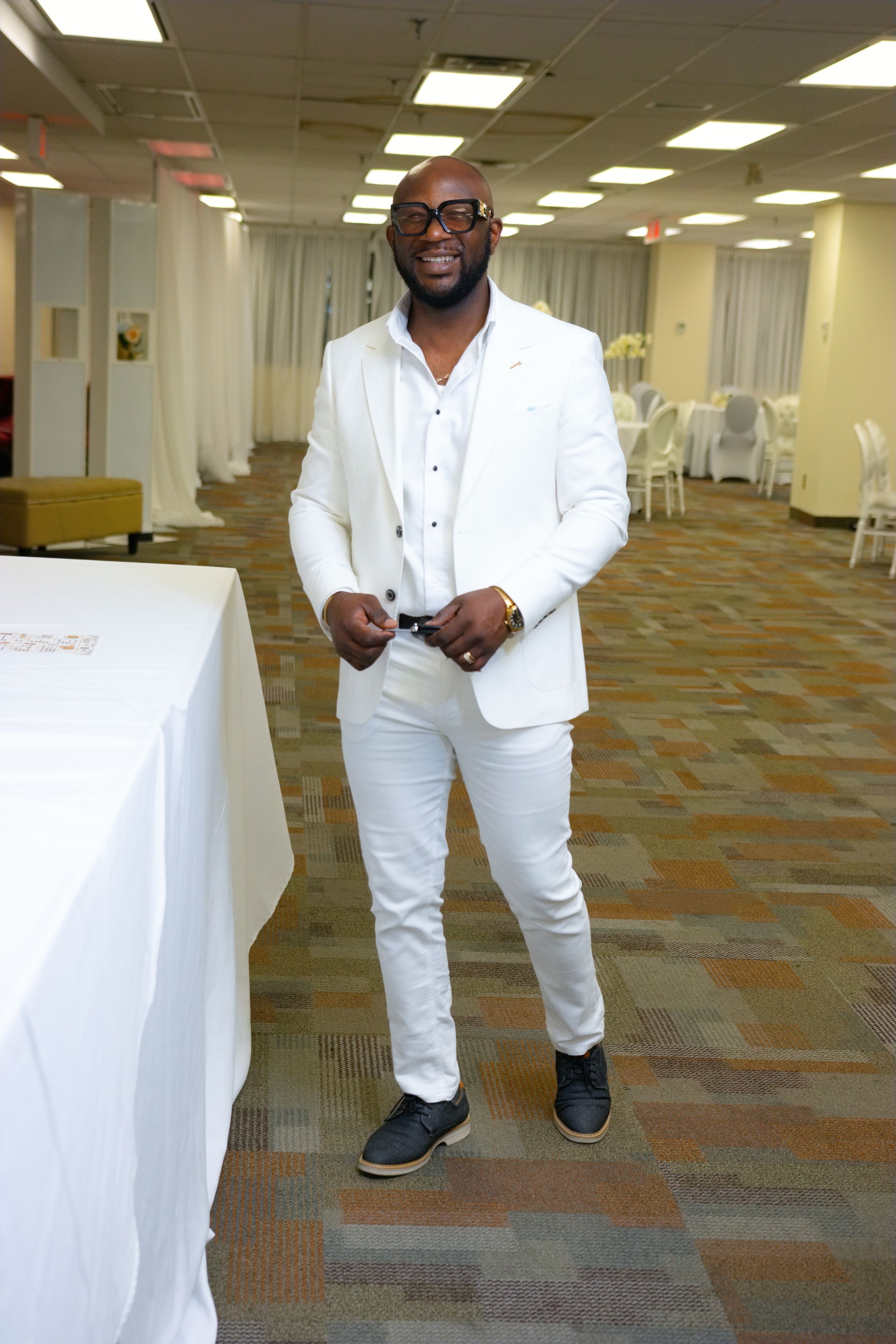 Stylish man in all-white suit and glasses posing confidently in an elegant event hall with patterned carpet.