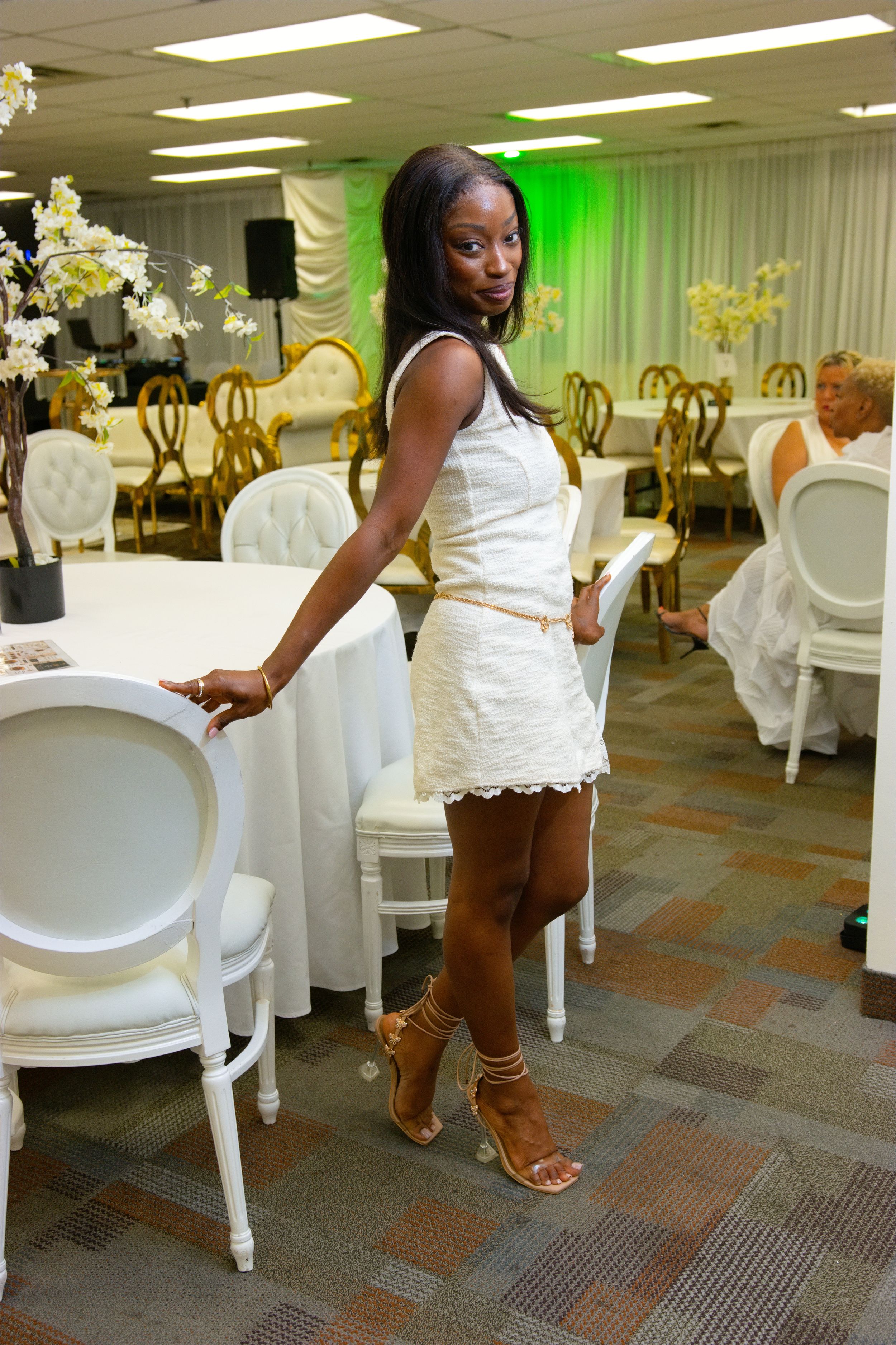 Elegant woman in white lace mini dress and gold heels poses in decorated event venue with white chairs and tables.