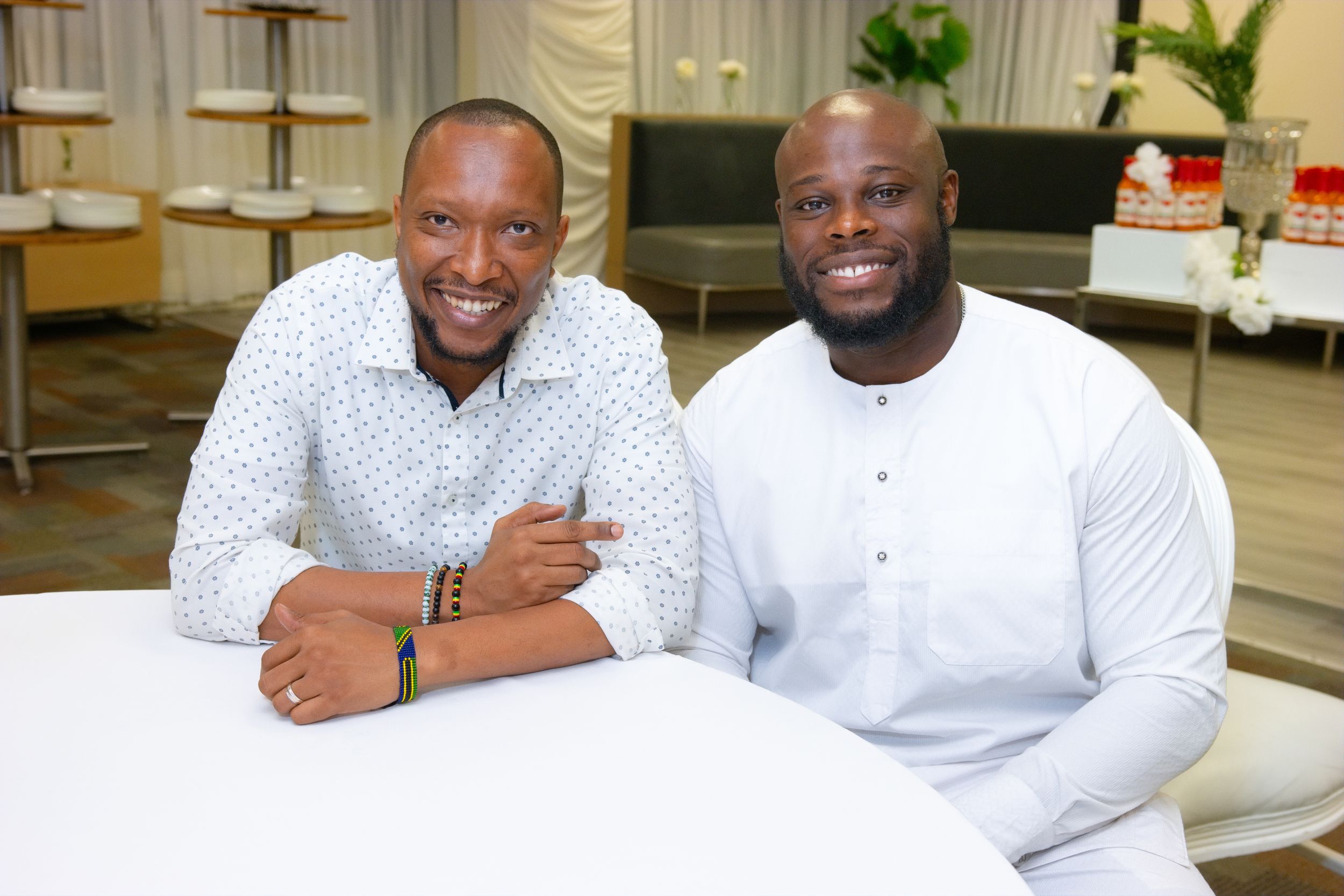 Two smiling men in white shirts pose together at an indoor event with elegant table settings in the background.