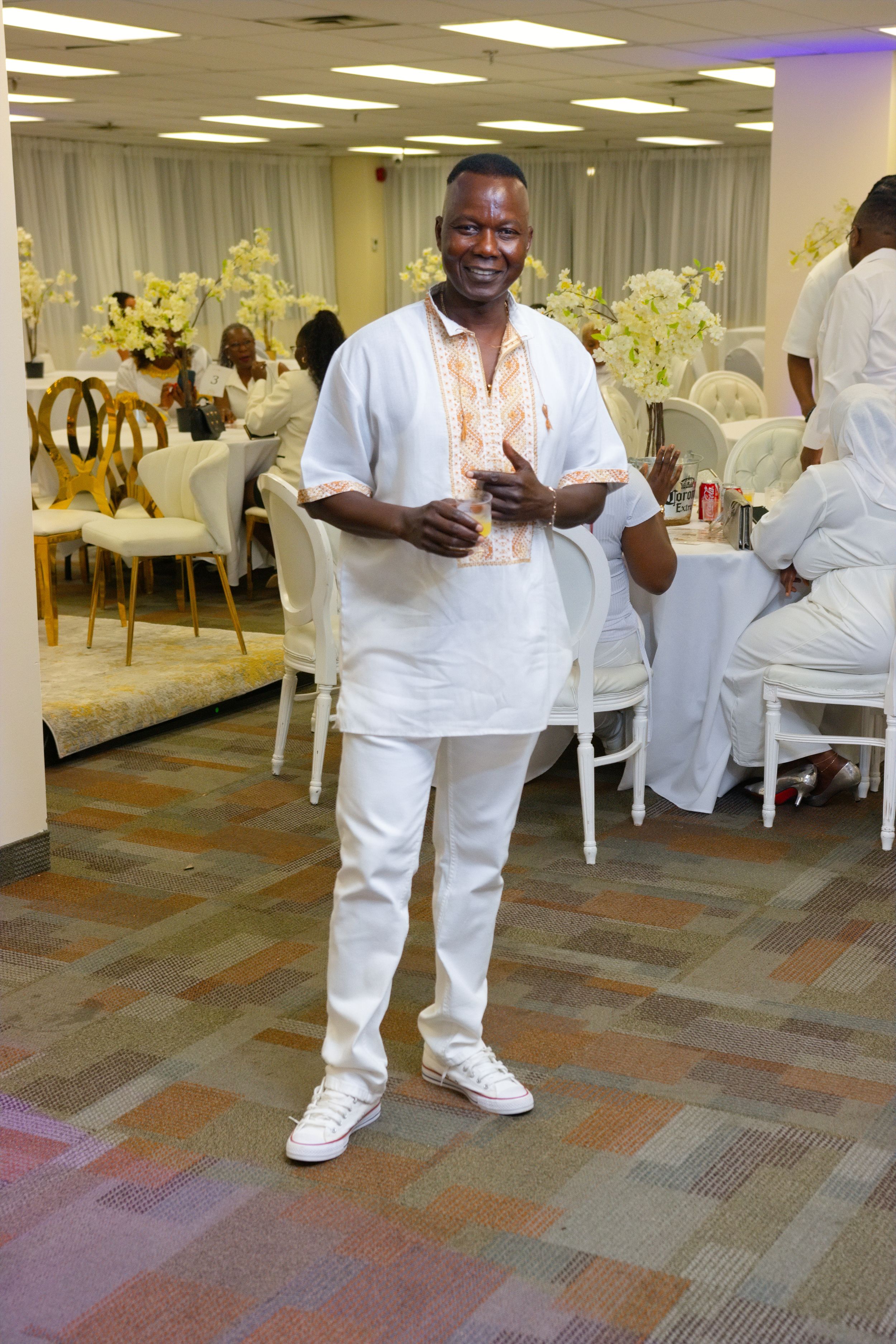 Man in white outfit smiling at an elegant event with white chairs and floral decorations.