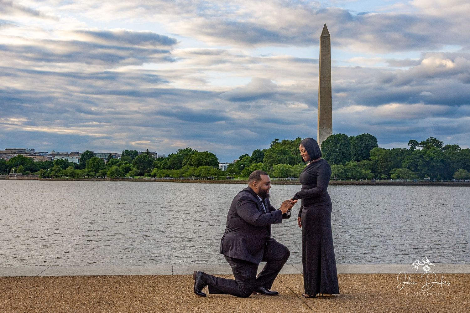 Man proposing to woman on one knee by the Tidal Basin with Washington Monument backdrop at dusk.