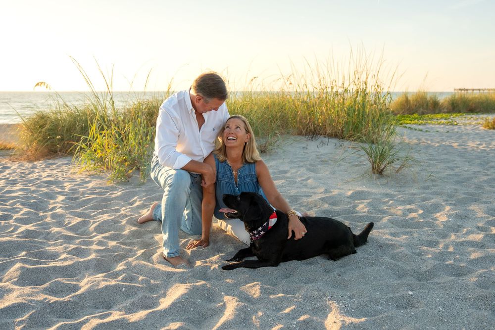 Couple relaxing on sandy beach with black Labrador dog near ocean dunes at golden hour sunset.