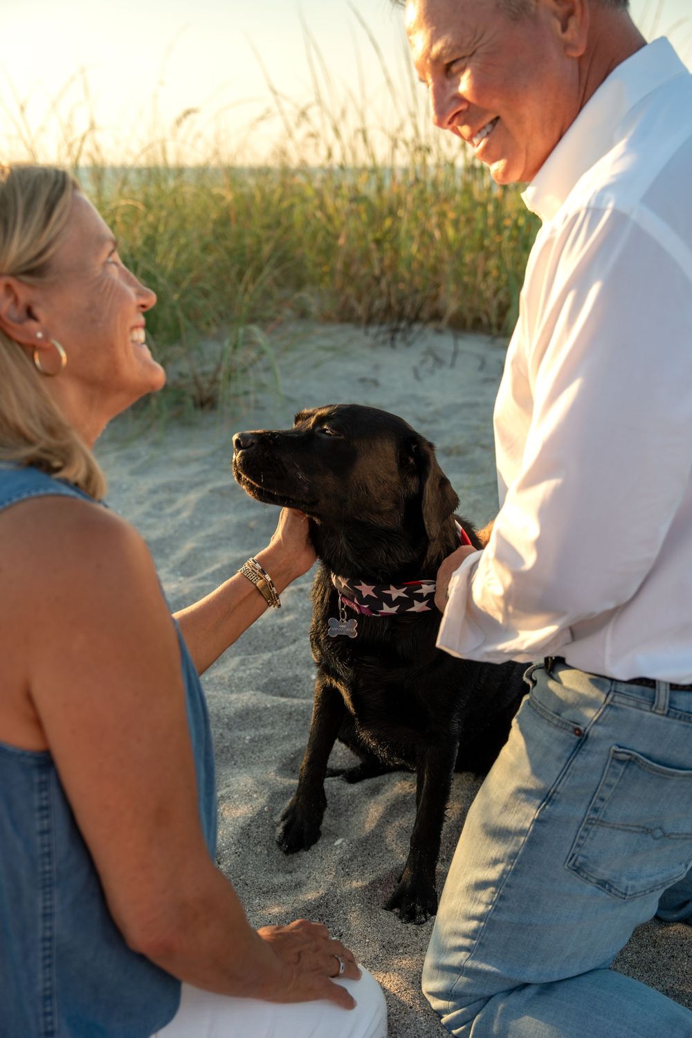 Smiling senior couple with black Labrador dog on sandy beach near tall grass at sunset.