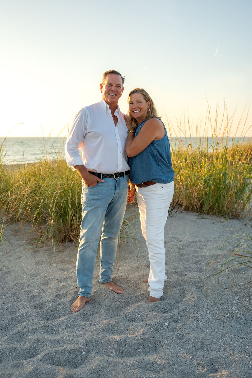 Smiling couple posing on sandy beach at sunset, man in white shirt and jeans, woman in blue top and white pants.