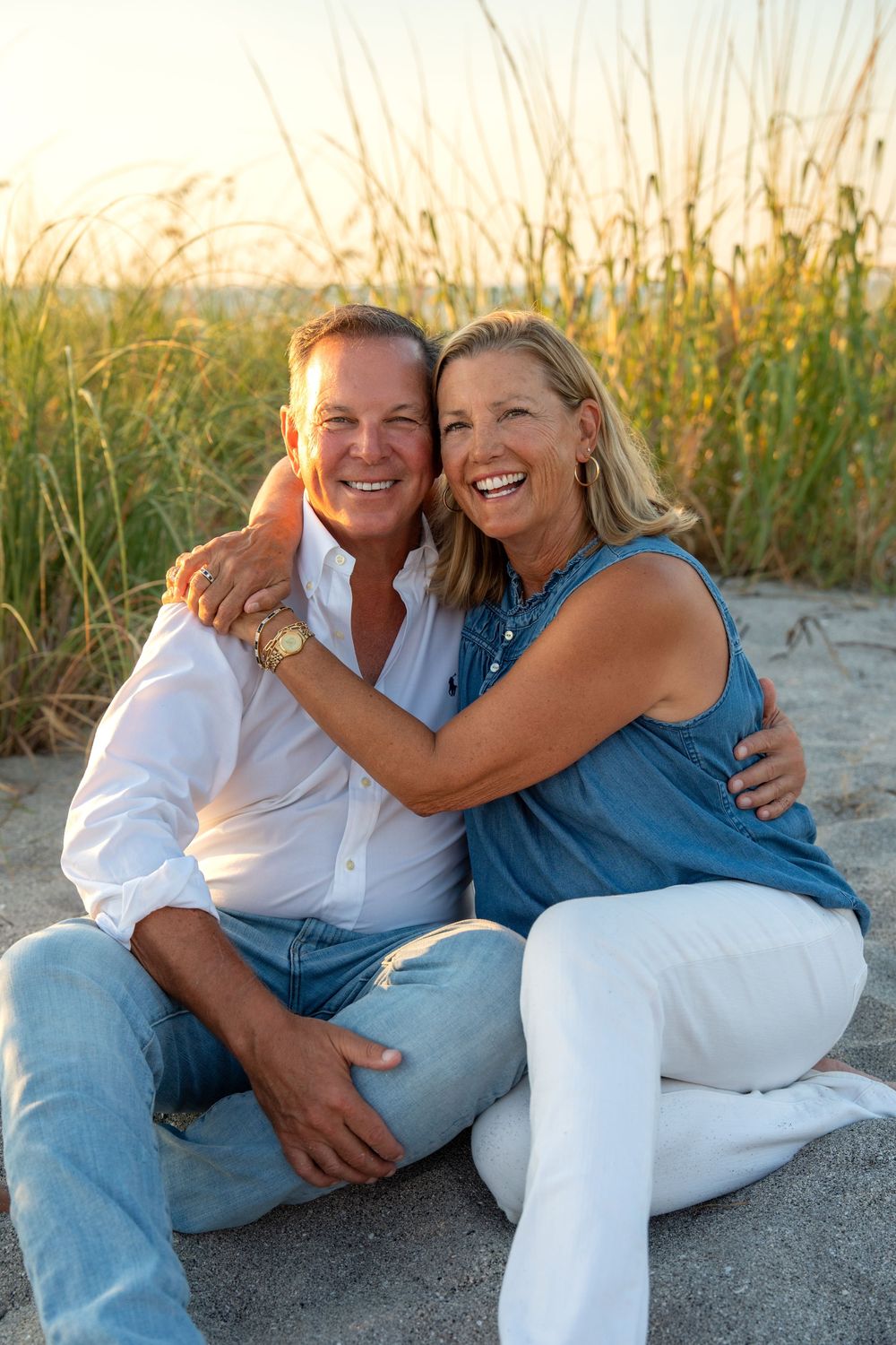 Happy middle-aged couple embracing and smiling on sandy beach at golden hour with tall grass in background.