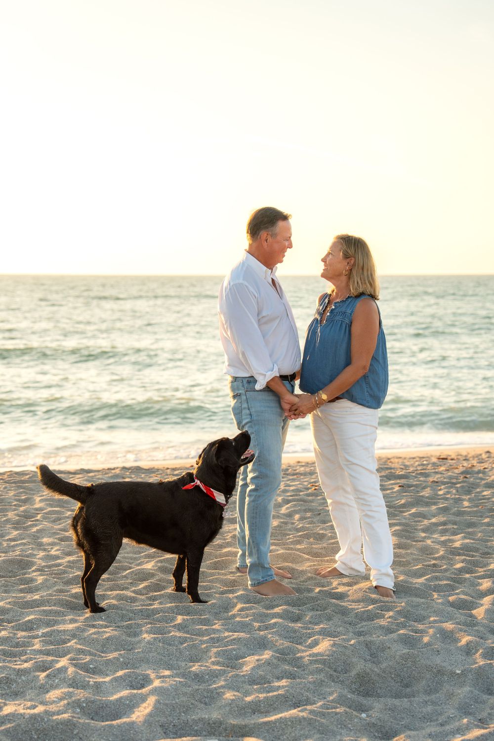 Couple holding hands on sandy beach at sunset with black Labrador dog, ocean waves in background.