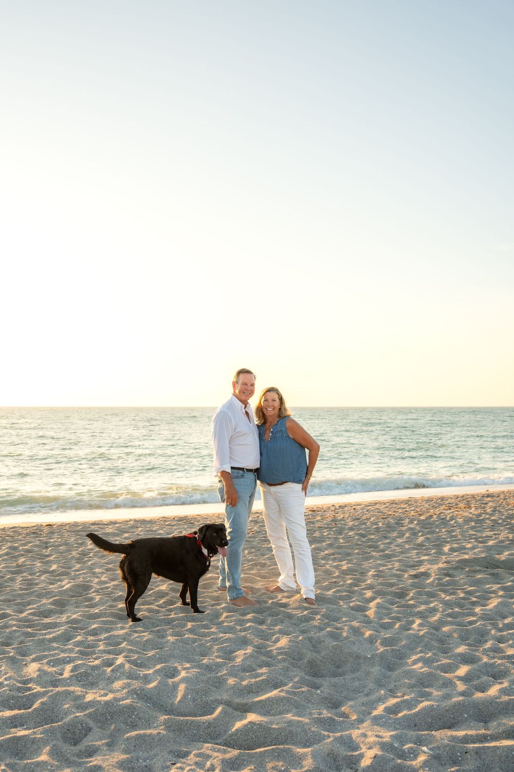 Happy couple walks black Labrador Retriever on sunny beach with calm ocean waves at sunset.
