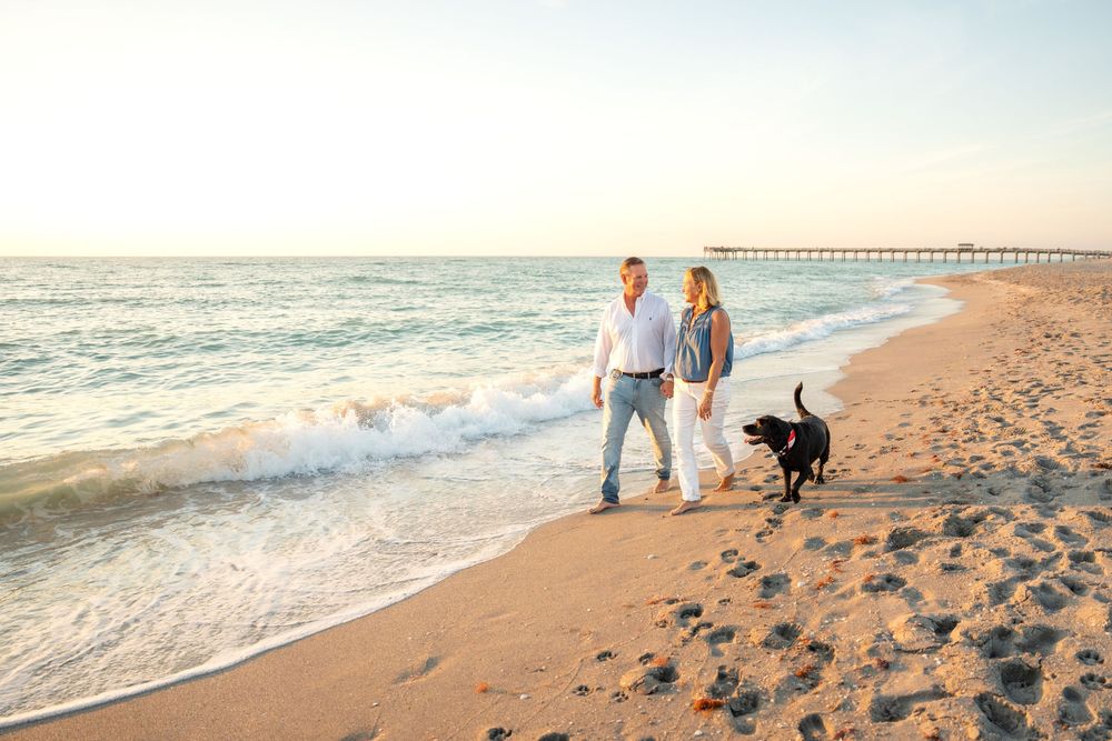 Couple walking black dog on sandy beach at sunset near pier.