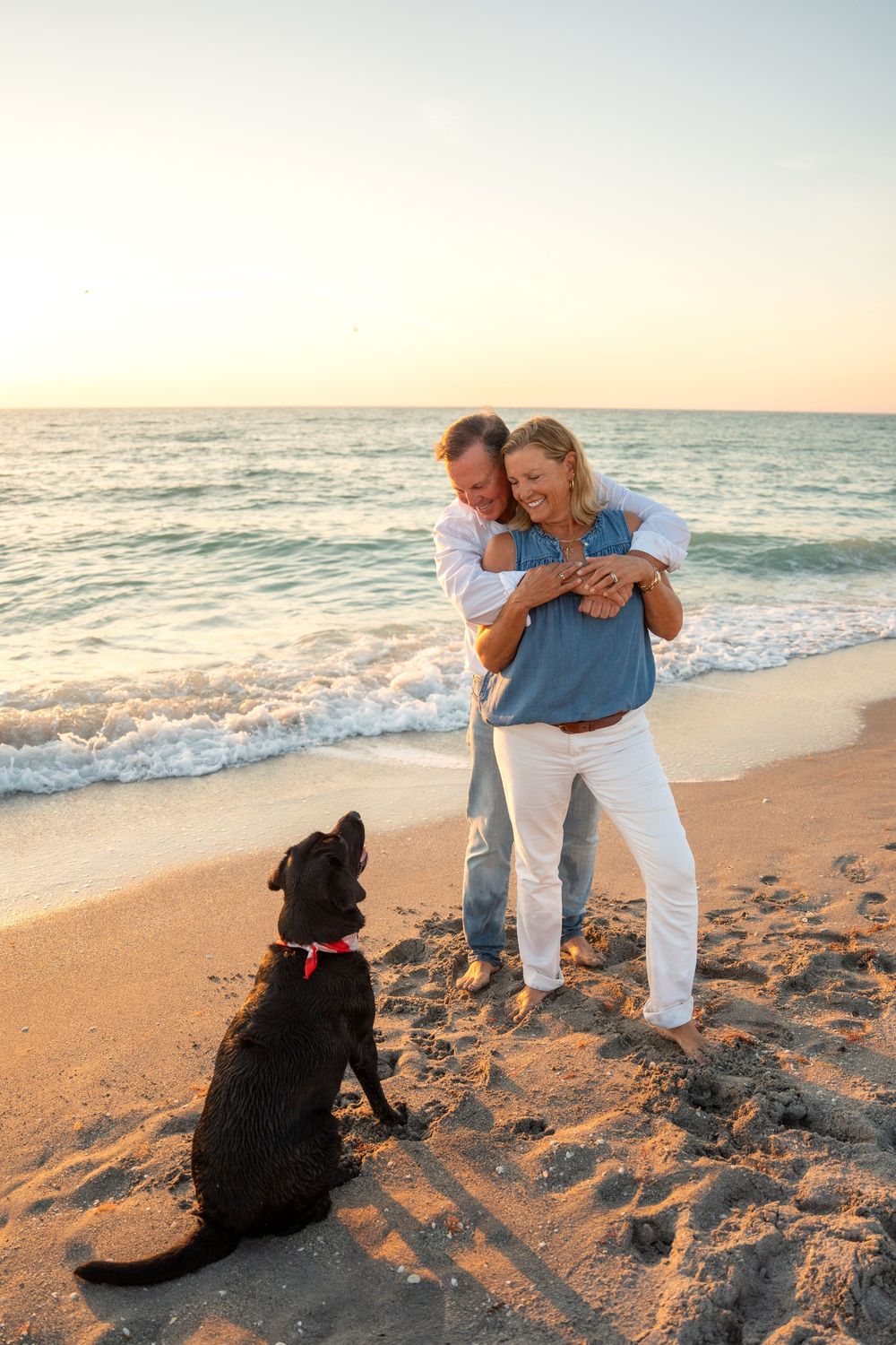 Man embraces woman from behind on beach at sunset as black Labrador watches adoringly.