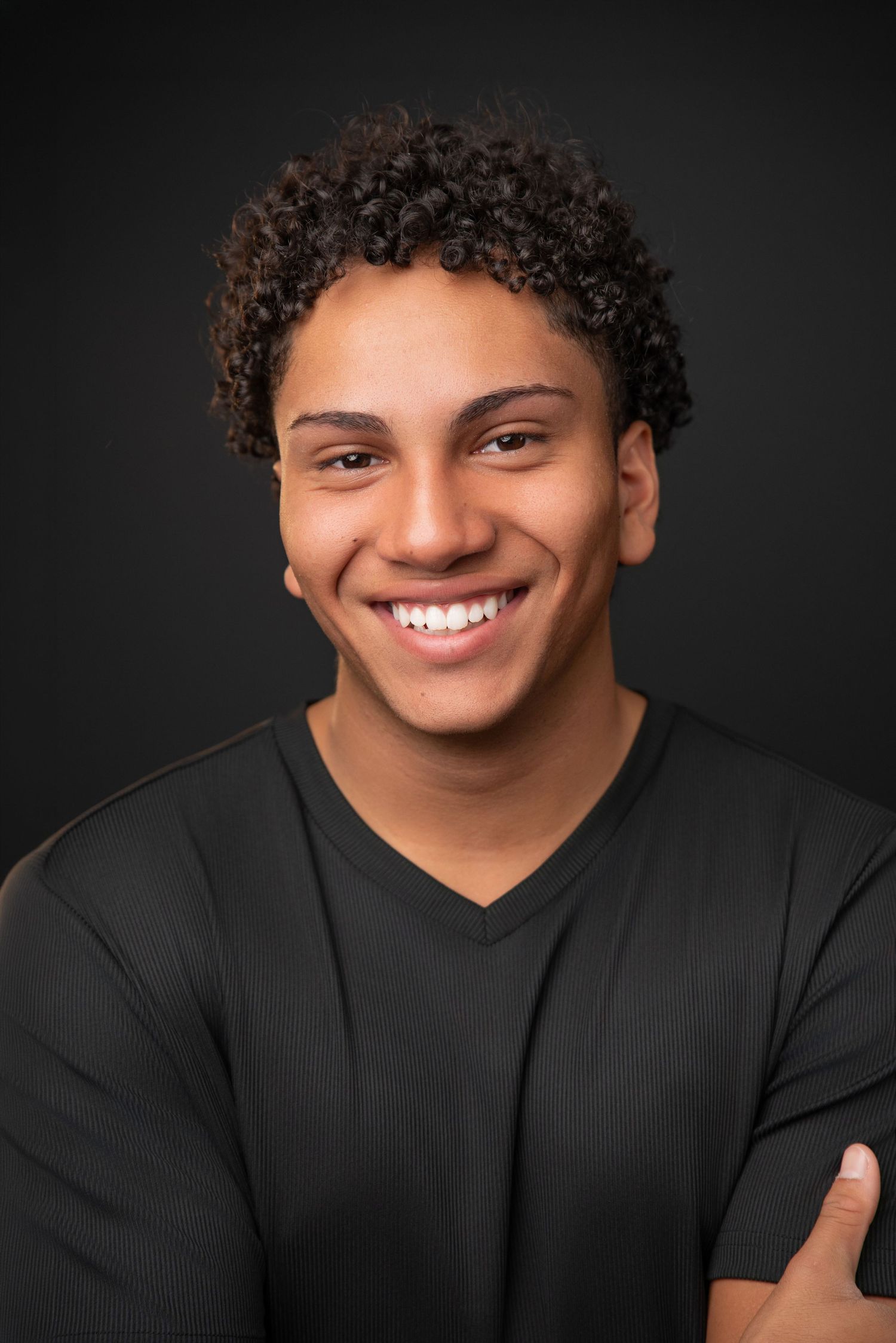 Young man with curly hair smiling confidently, wearing a black v-neck shirt against a dark background.
