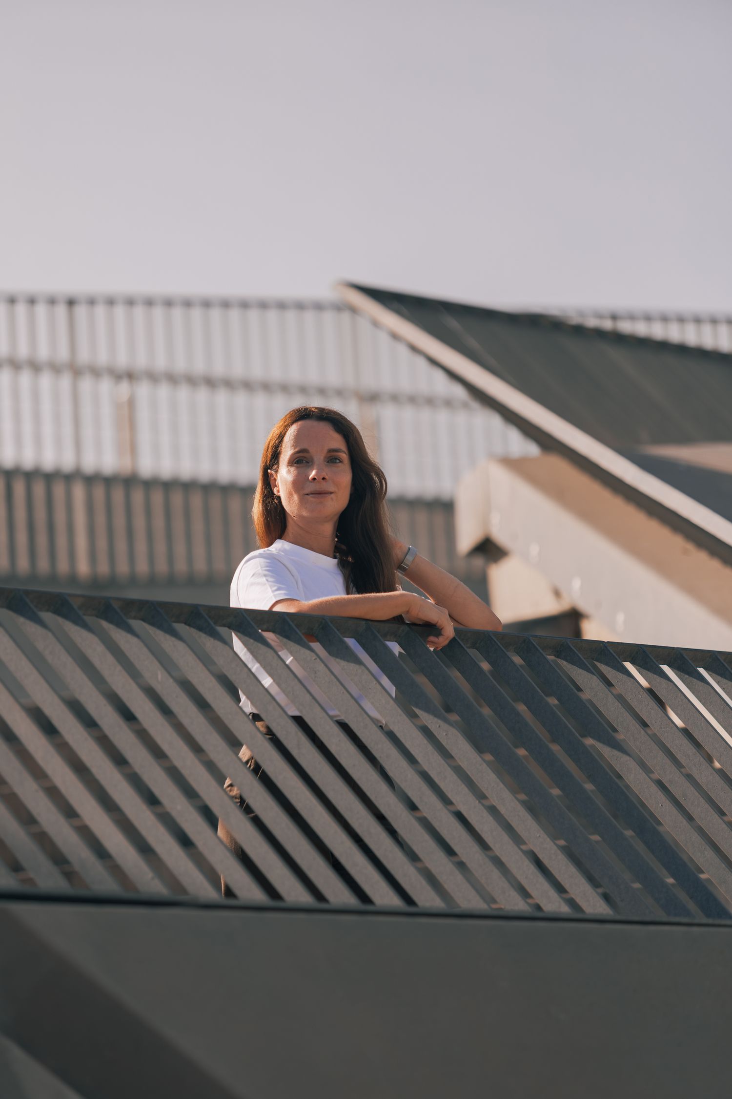 Woman in white t-shirt leaning on metal railing outdoors, looking thoughtfully into the distance against a clear sky.