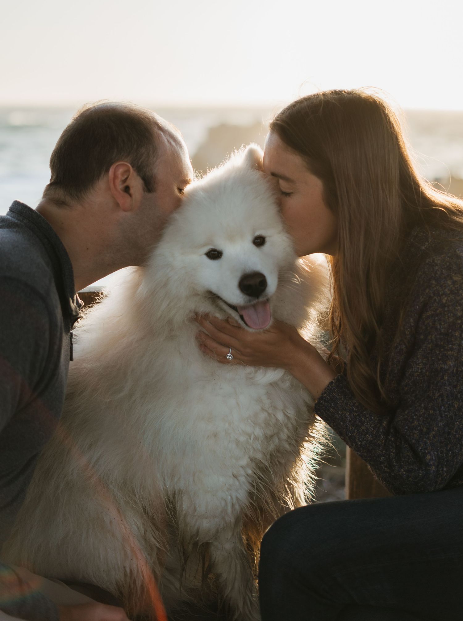 Couple kissing their fluffy white Samoyed dog on the cheeks outdoors near water at sunset.