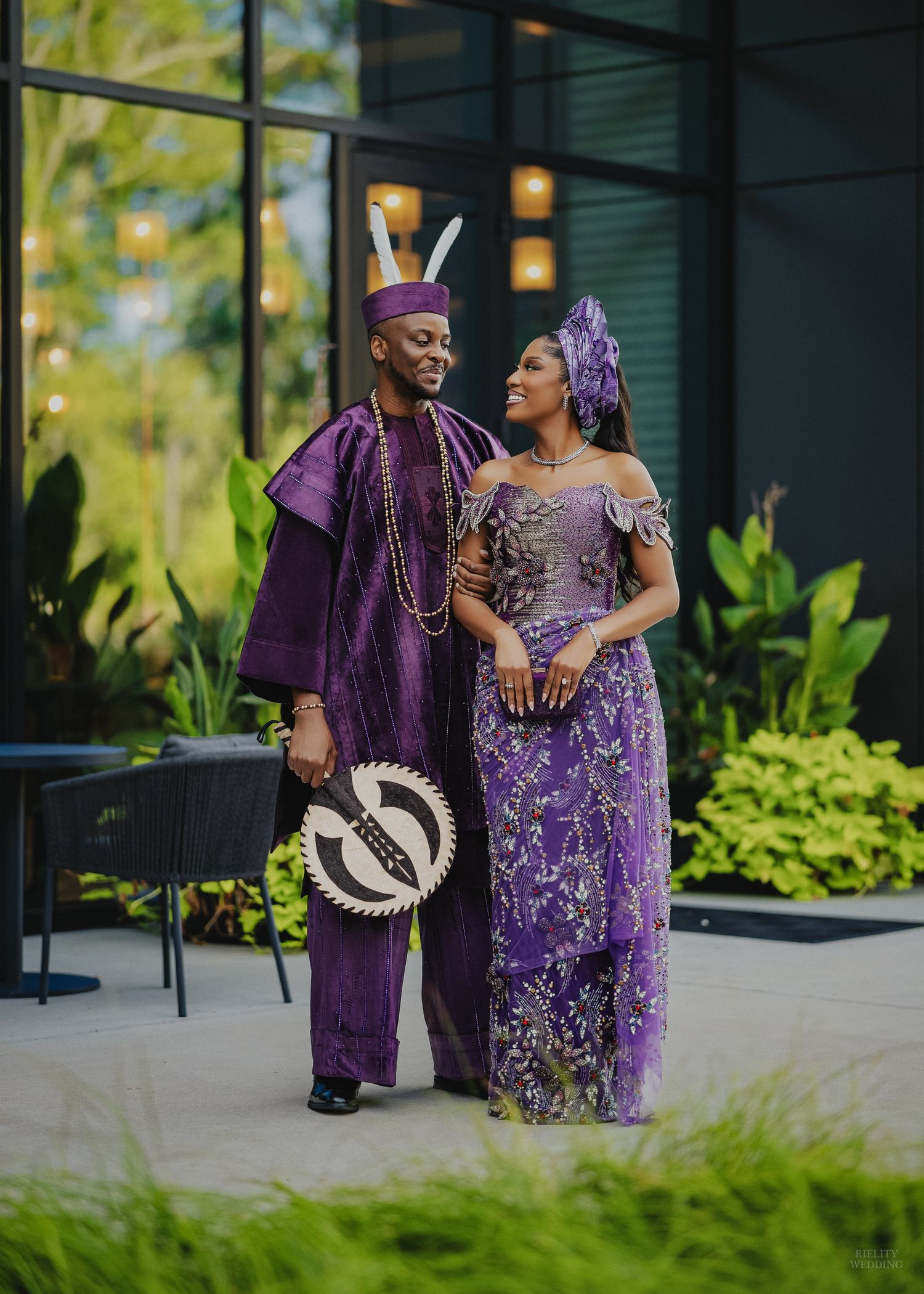 Couple in matching purple traditional African attire posing outdoors, man in agbada with beaded necklace, woman in lace gown.