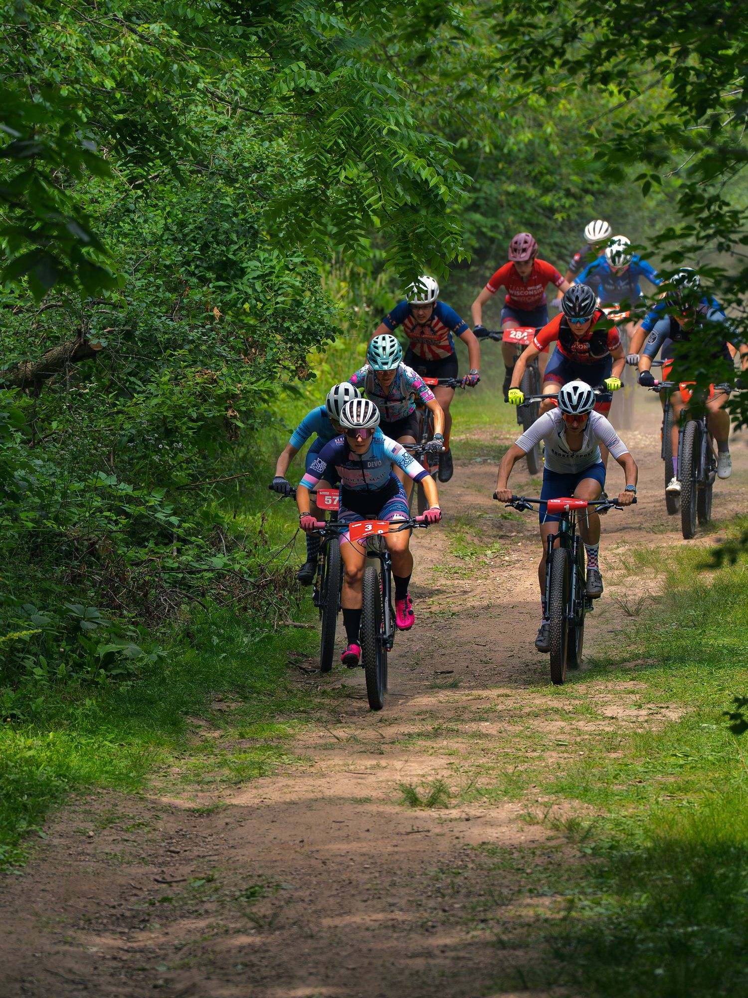 A pack of mountain bike racers climbs a narrow dirt singletrack lined with dense green summer trees, front riders leading the field uphill.