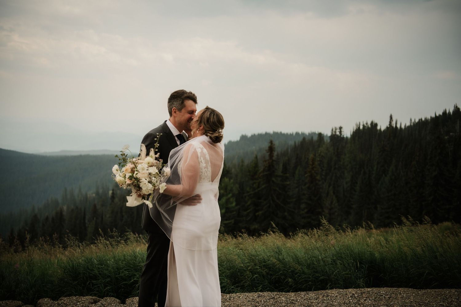 Bride and groom share a romantic kiss on a mountain overlook surrounded by evergreen forests on their wedding day.