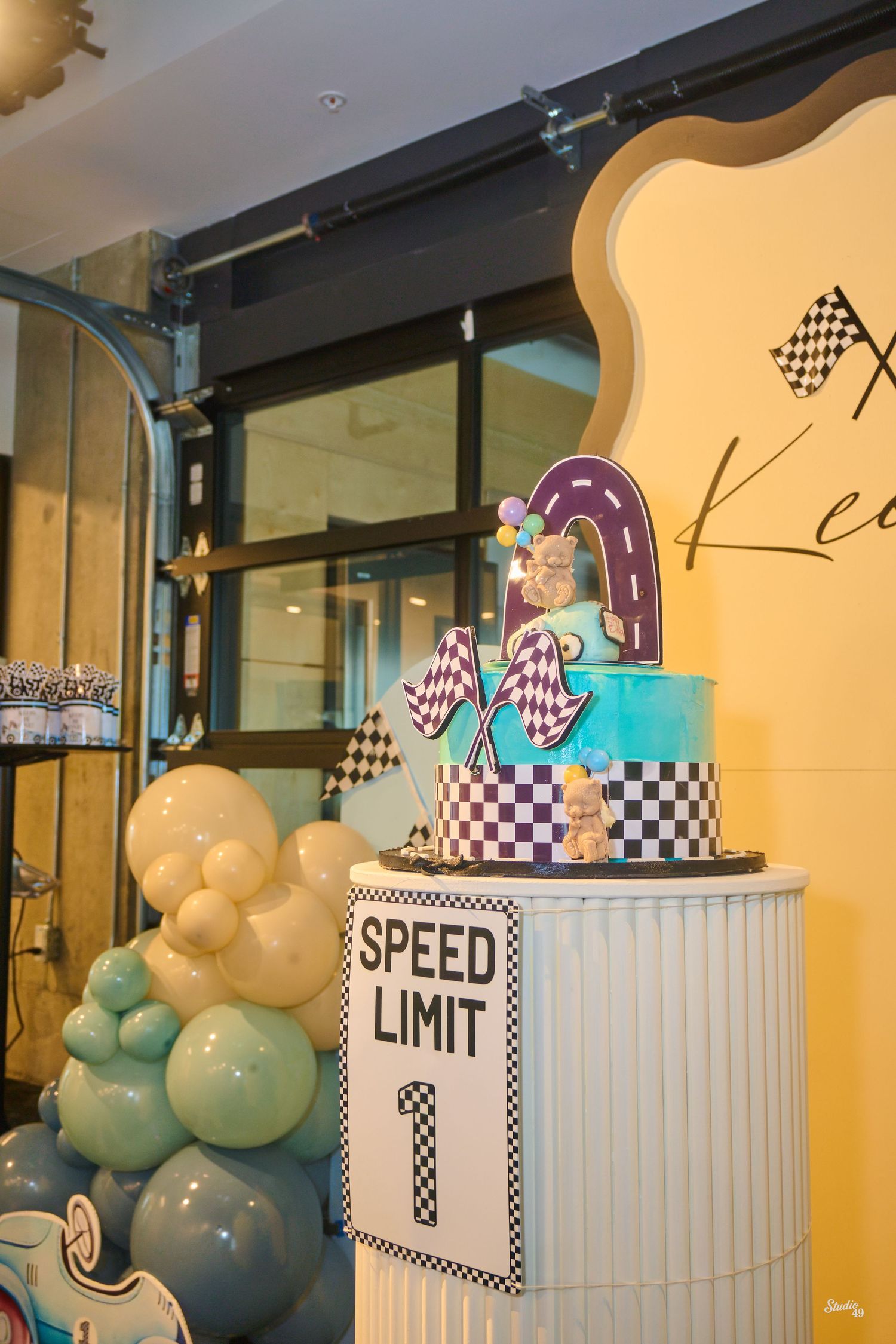 A race car themed first birthday cake display with checkered decor and Speed Limit 1 sign surrounded by blue balloons.