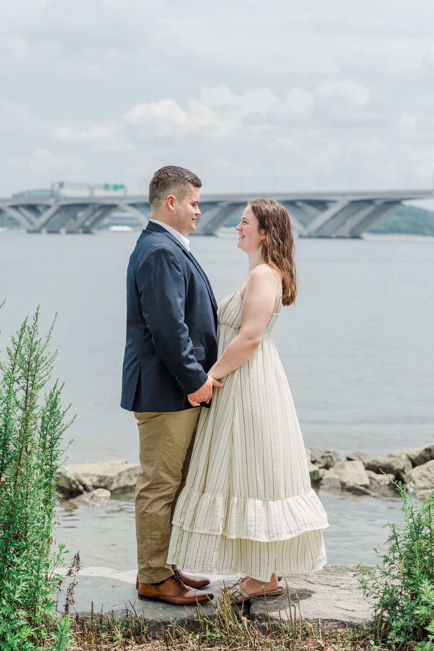 Couple holding hands and smiling at each other by a waterfront with a bridge in the background on a cloudy day.