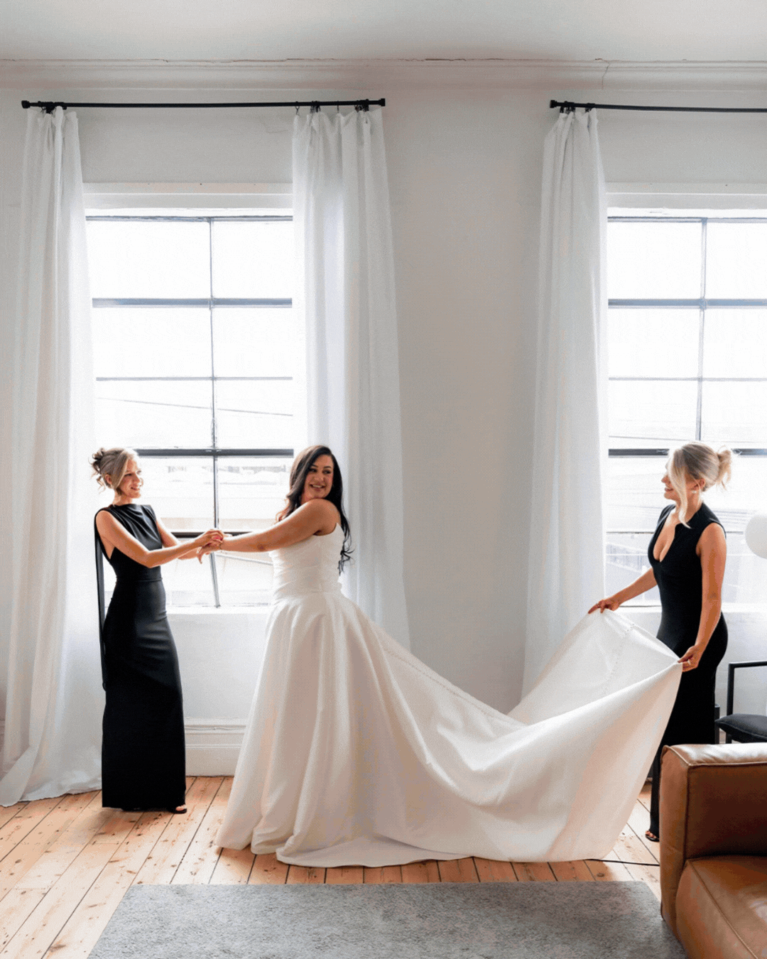 Bride in white gown being helped by two bridesmaids in black dresses near bright windows on hardwood floors.