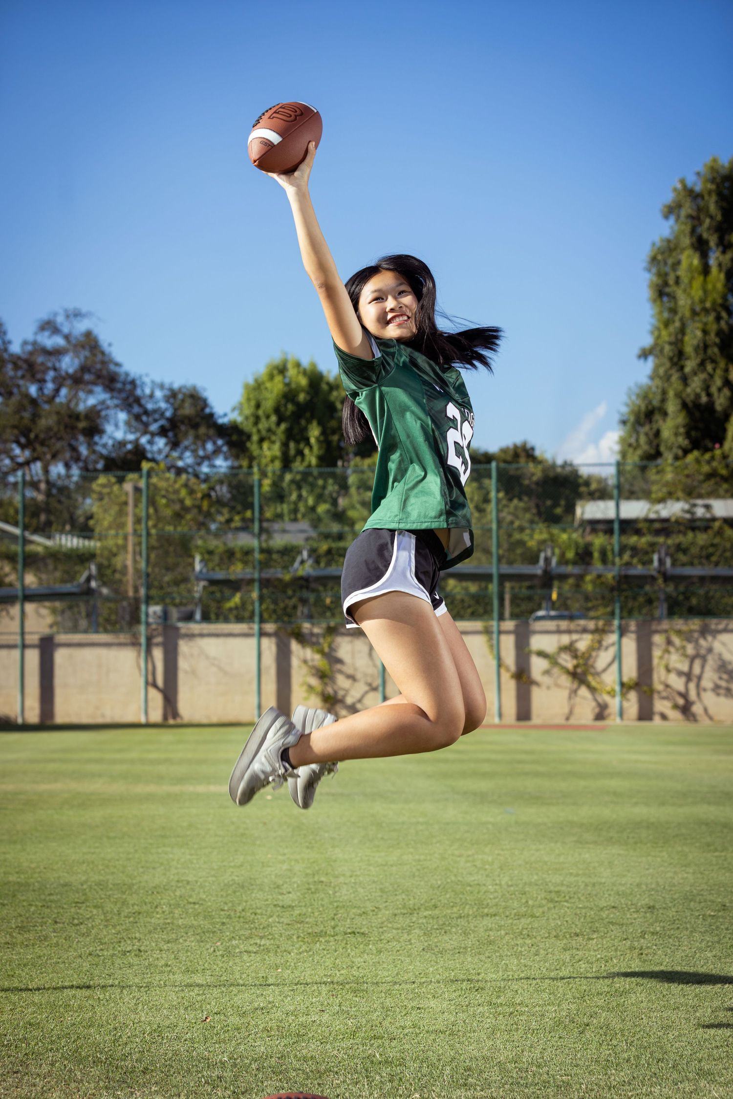 Young woman in green football jersey jumping mid-air while holding a football on a green field.