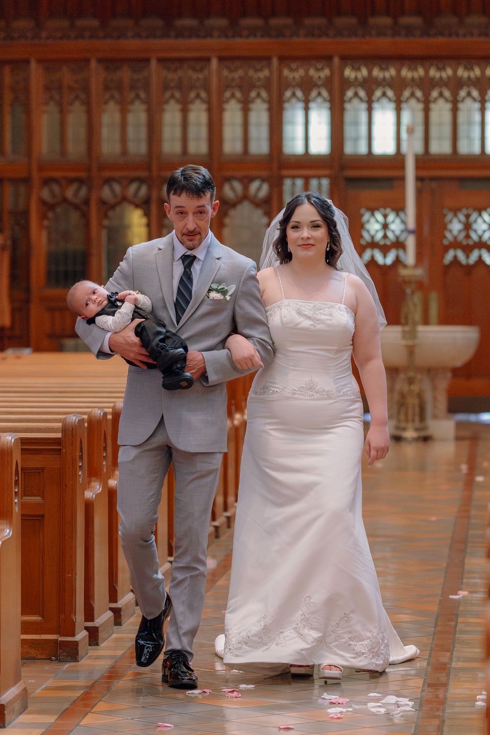 Bride and groom walk down church aisle holding newborn baby after wedding ceremony.