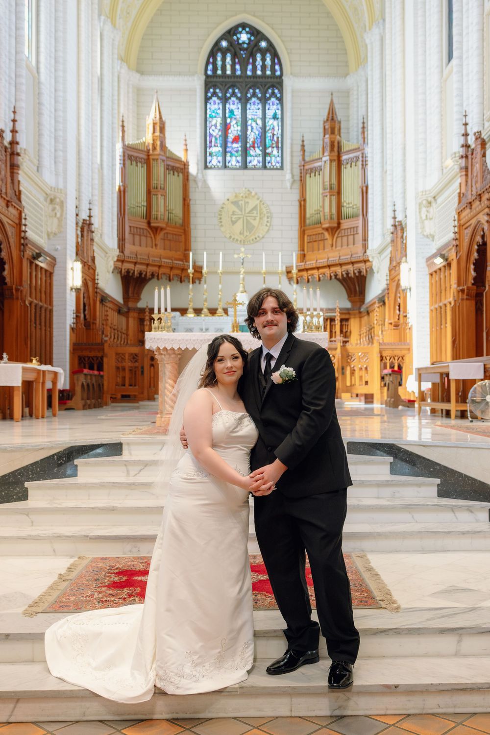 Bride and groom posing together at church altar, bride in white gown, groom in black suit, stained glass windows behind.