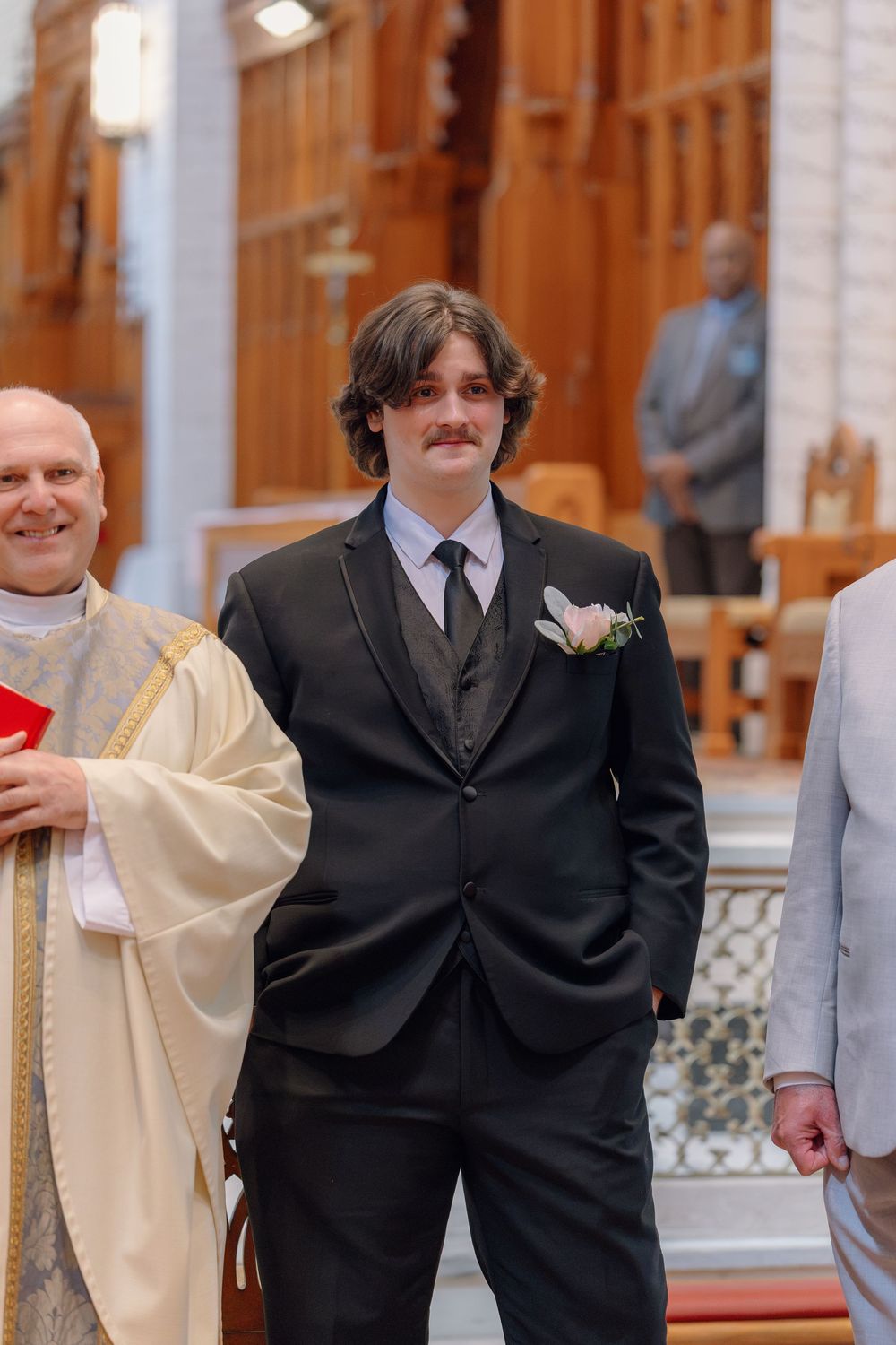 Young groom in black suit with boutonniere stands in church aisle beside smiling priest in ceremonial robes.