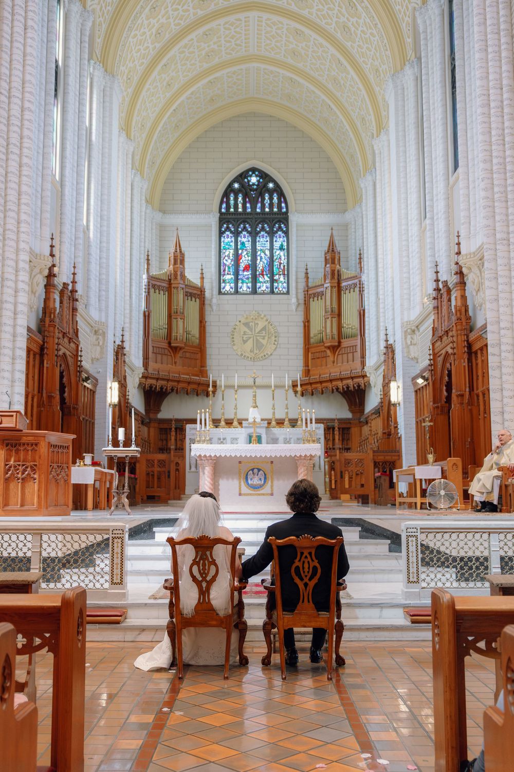 Bride and groom seated at church altar during wedding ceremony in ornate cathedral with stained glass windows.