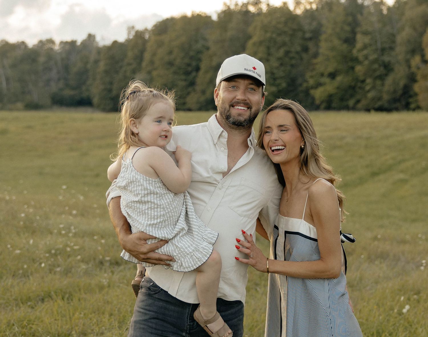Happy family of three posing in a green meadow, man holding young girl, woman in striped dress, trees in background.