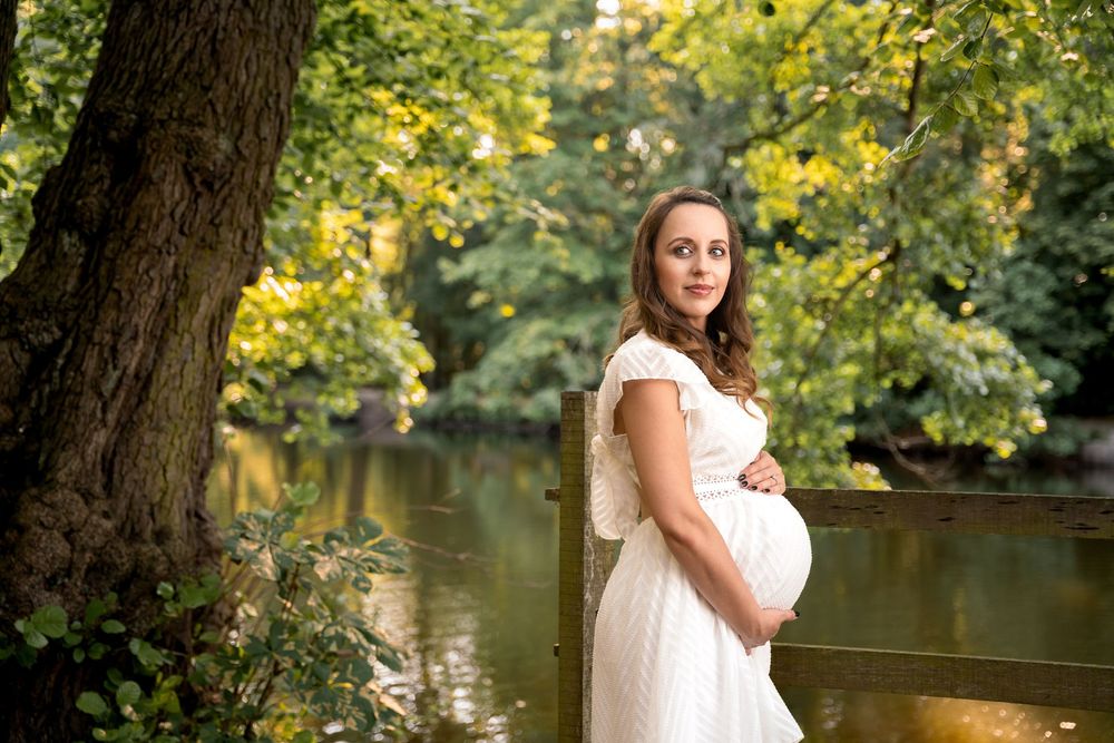 Pregnant woman in white lace dress holding belly outdoors near a serene river surrounded by lush green trees.