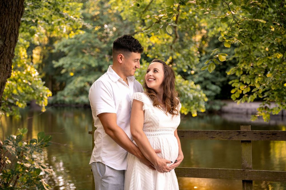 Couple embracing outdoors near a pond, woman in white dress cradling baby bump during maternity photo session.